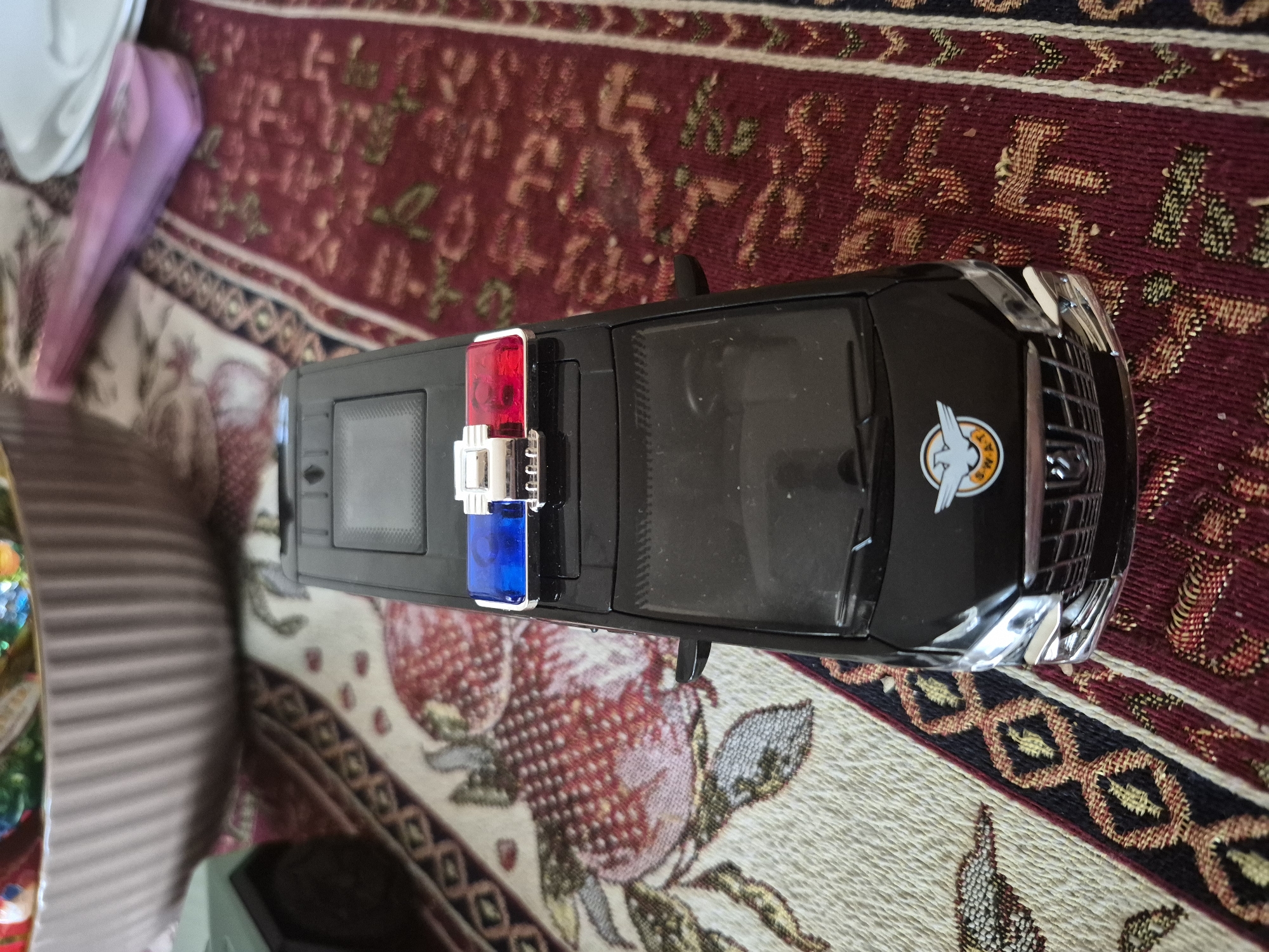 A black toy police vehicle, resembling an SUV, is prominently displayed diagonally across the frame, positioned on a richly patterned rug. The vehicle features distinct red and blue emergency lights on its roof and a chrome-colored front grille. On its side, near the front, a circular emblem shows a stylized white bird, possibly a falcon, within an orange circle. Below the bird, faint text is visible, appearing to start with letters like "ALLIAM" or "WILLAM."
The vehicle rests on a multi-colored rug with intricate designs. The dominant color is a deep red, adorned with traditional patterns in lighter reds, greens, and browns, which could be floral or abstract motifs. A lighter section of the rug, visible beneath and to the left of the car, has a beige background with larger red patterns that resemble strawberries. A decorative border featuring dark blue, cream, and gold geometric shapes is also visible. In the blurred background, hints of other objects include a light-colored fabric and a pink or purple item to the upper left, and a striped, light brown object to the lower left.
The scene is static, depicting objects only, with no people or activity. The traditional rug patterns are consistent with styles found in regions like Yerevan, Armenia. There are no discernible time-of-day or weather cues, nor any explicit emotions conveyed.