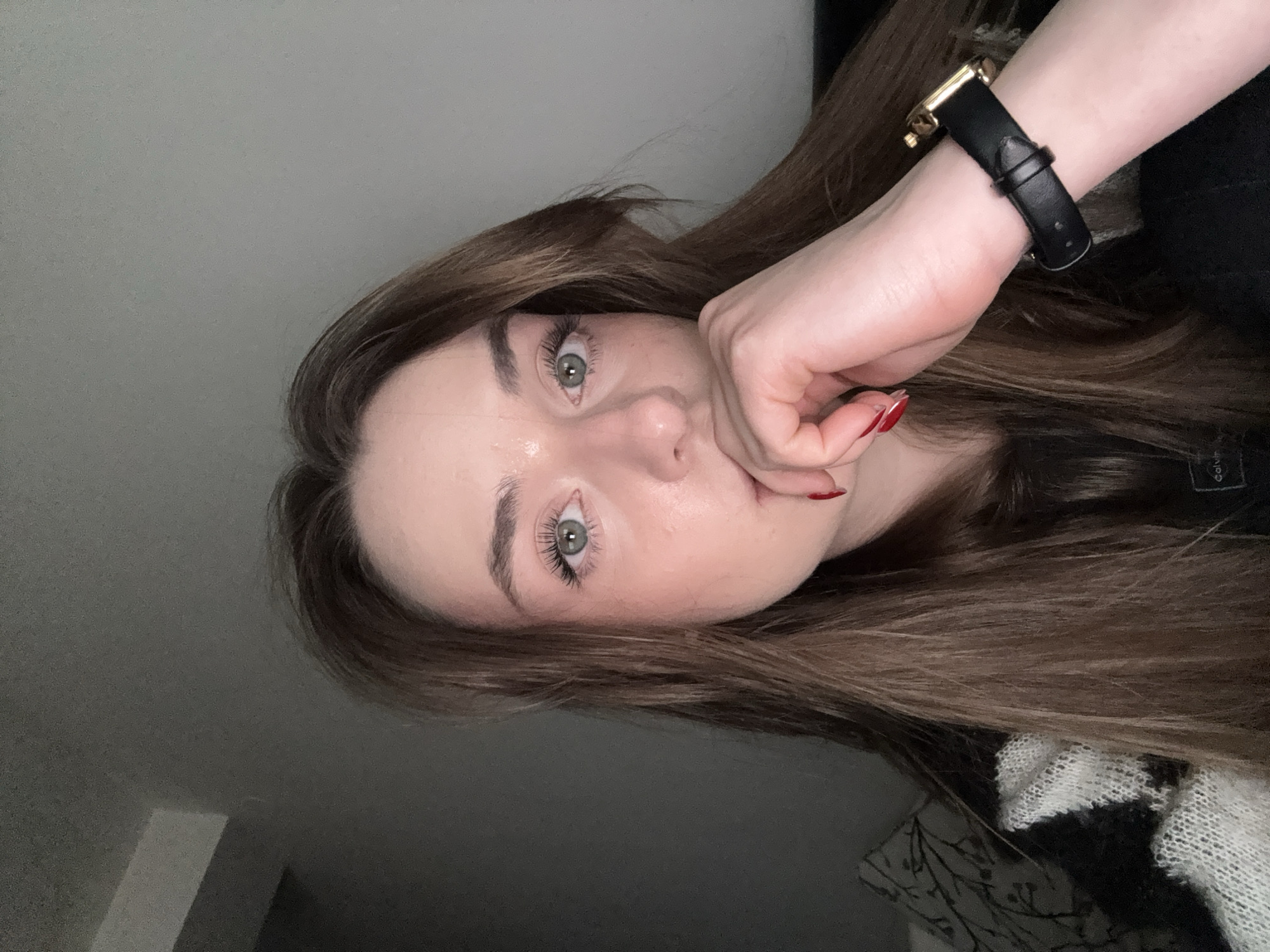 A young woman with fair skin, light brown hair, and green eyes poses for a selfie. She is wearing a black wristwatch with a gold buckle on her left wrist. Her hand is near her mouth, with red nail polish visible on her fingernails. Her hair is long and styled, and she appears to be wearing light makeup. She is wearing a black and white patterned garment. The background is a neutral, greyish tone. The photo does not appear to be taken in the city of Kragujevac, Serbia. There are no elements indicating the location.