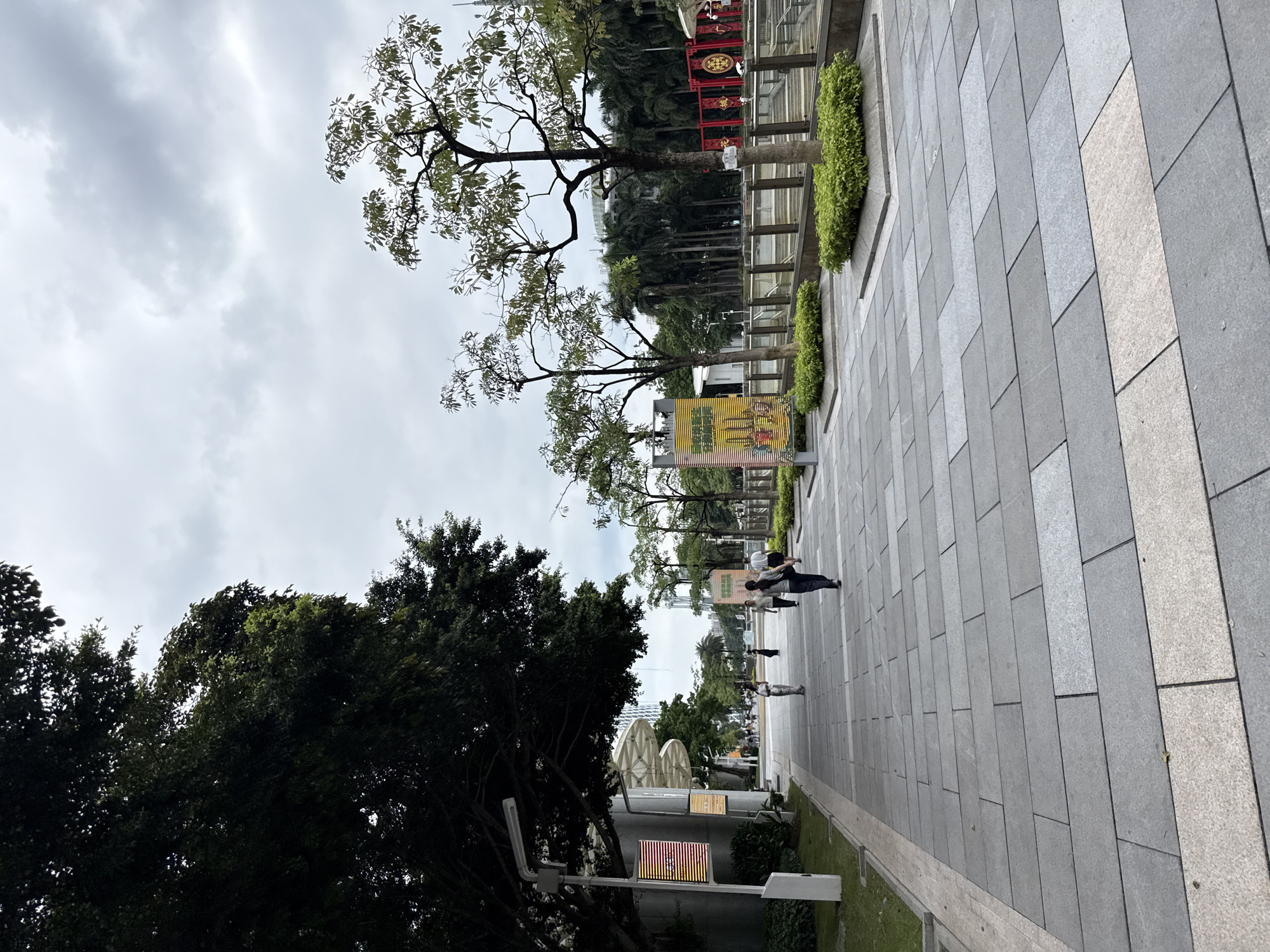 The image depicts a pedestrian walkway in Tianhe District, China, under a cloudy sky. The walkway is paved with large, grey rectangular tiles with a lighter border. To the left, a large tree with dark green foliage shades a section of the path, next to a building with a curved facade and a light-colored awning. A lamppost with two illuminated displays stands near the tree. Several people are walking along the path, some dressed casually. In the background, smaller trees with lighter green leaves are planted in square, raised beds filled with manicured green shrubs. Beyond these, a row of palm trees and other vegetation forms a backdrop, with modern buildings visible in the distance. A fenced area with red decorative elements and signs is also visible in the far background.