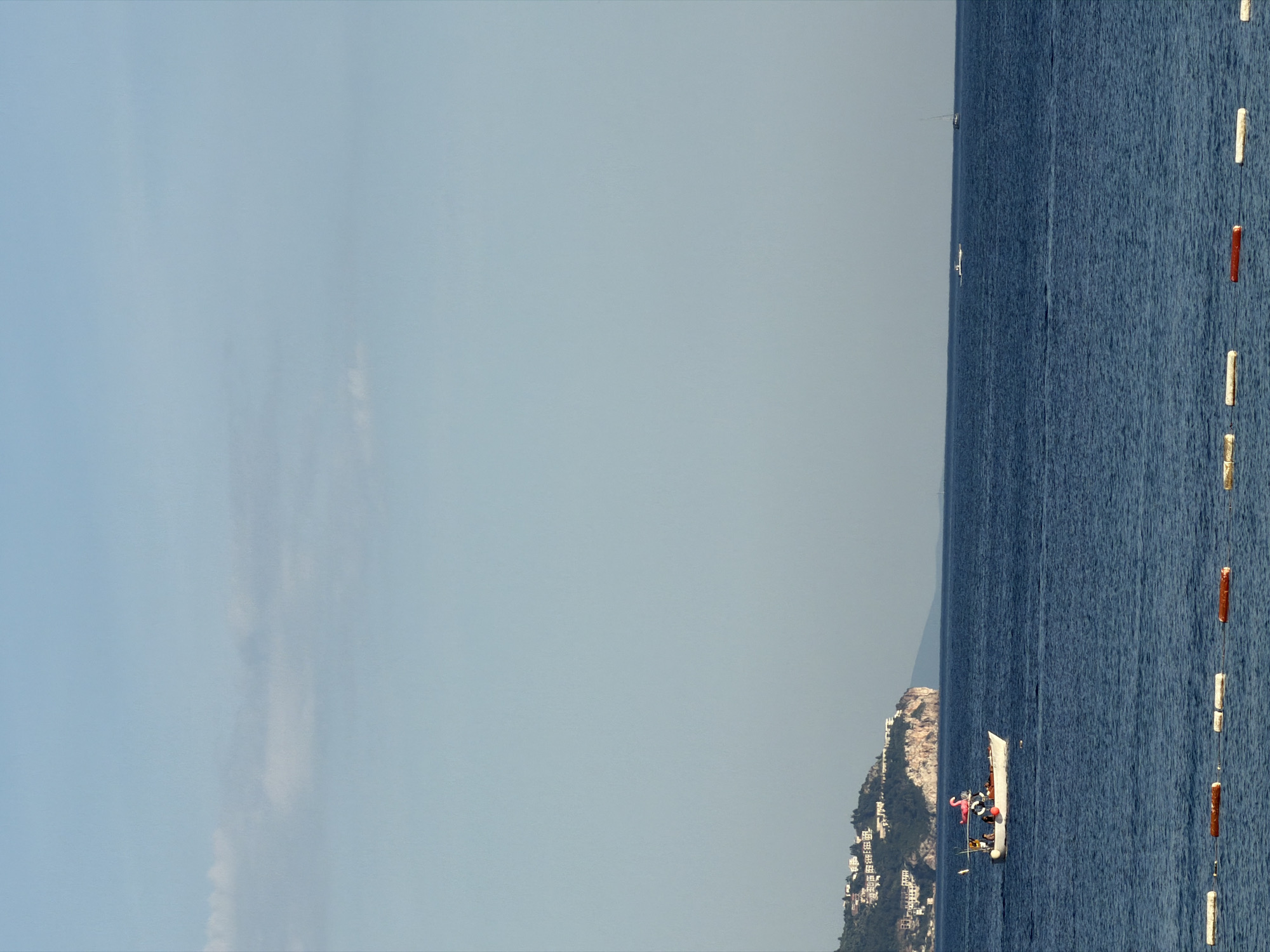 The image depicts a seascape with a boat and a distant coastline. The right side of the image is dominated by the dark blue sea, where a line of white and orange buoys stretches vertically. In the middle, a white boat with several figures, including one wearing a pink inflatable flamingo costume, is visible. The coastline in the background features a hill with a dense concentration of white buildings. The sky is a pale blue, occupying the left side of the image, and is almost cloudless. While Budva, Montenegro is mentioned, it is impossible to confirm this as the specific location without additional information.