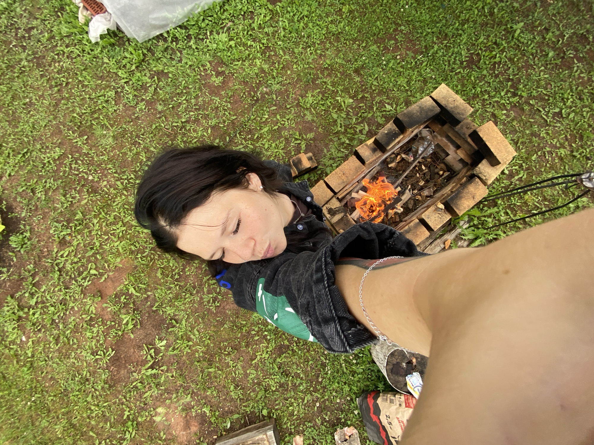 This is an overhead shot of a person lying on a grassy area near a brick fire pit. The person has dark hair and is wearing a dark denim jacket with a green graphic on the sleeve. A silver bracelet is visible on their arm, and a tattoo can be seen on their bicep. The fire pit contains burning wood, casting a warm orange glow.
The ground is covered in short green grass and patches of dirt. To the left of the fire pit, there is a white plastic sheet and some white material with red brick patterns visible. In the bottom right corner of the image, a bag of charcoal briquettes is partially visible with text that appears to read "2,5 kg". The overall scene suggests an outdoor setting, possibly a backyard or campsite. The lighting indicates it might be daytime, with overcast conditions contributing to the soft light. The person's relaxed posture and closed eyes could suggest a moment of rest or contemplation by the fire. The location is identified as Foki, Russia.