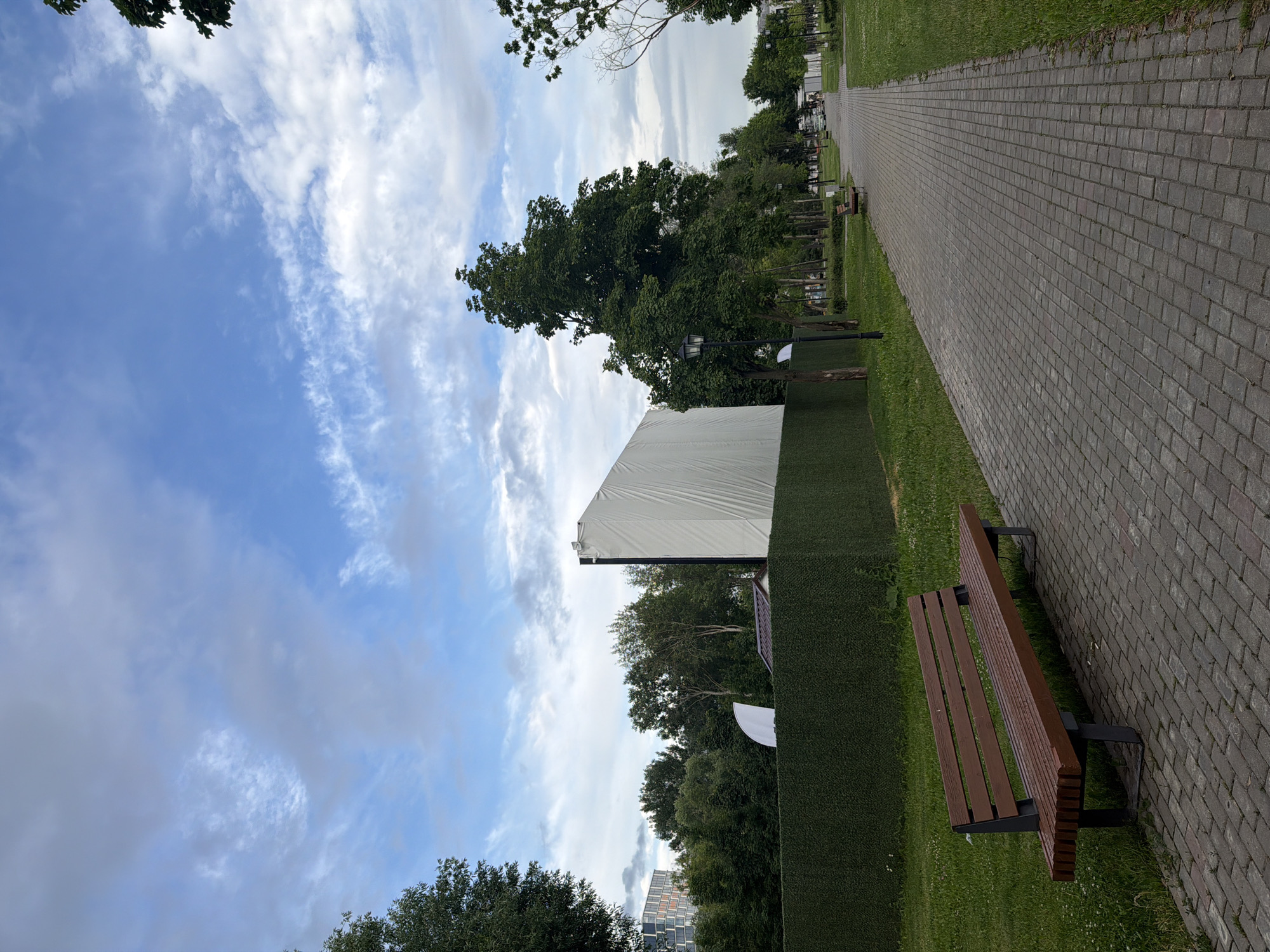 The image depicts an outdoor scene in what appears to be a park or public space. In the foreground, a brown wooden bench with dark metal legs sits on a paved walkway made of gray rectangular stones. To the left of the bench is a grassy area. The walkway extends into the distance, lined by more grass and trees on either side. In the middle ground, there is a tall, green hedge, and behind it, a large, white, rectangular screen is visible, suggesting it might be an outdoor movie screen. A tall, dark lamppost stands in front of the hedge, near the screen. Beyond the hedge and screen, there are more trees and some buildings in the far background, including a modern glass building on the left. The sky above is partly cloudy with patches of blue sky and white clouds. The overall impression is a serene, possibly urban park setting.