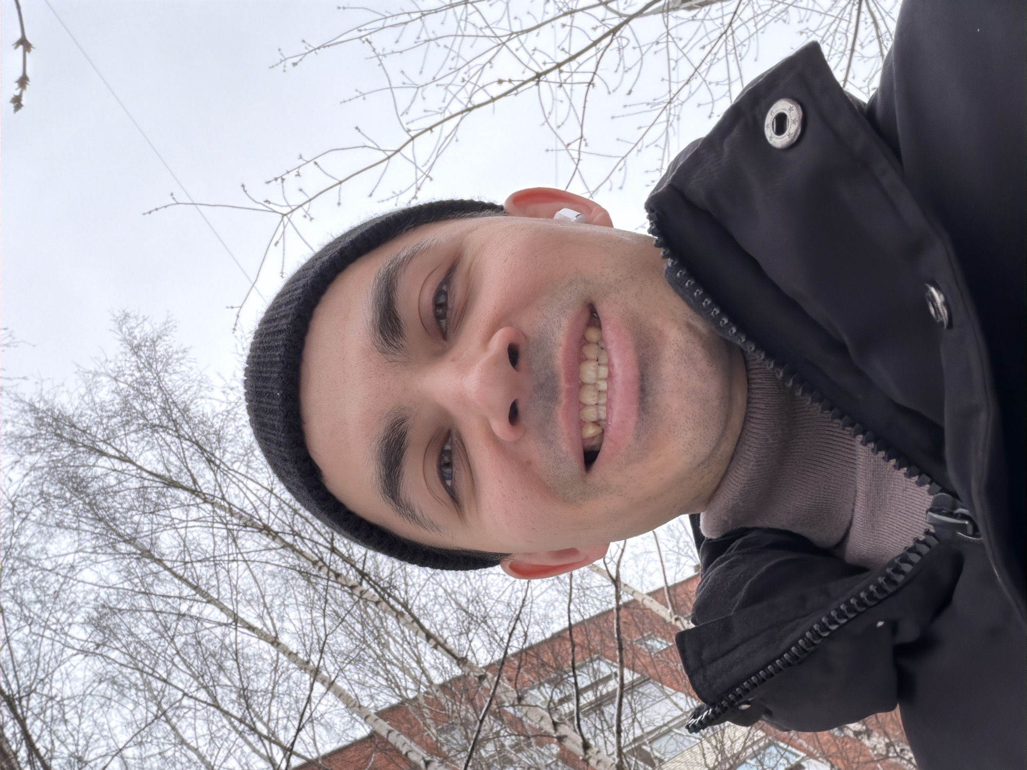 A close-up, low-angle selfie of a man with medium-brown skin, wearing a black beanie, black winter coat, and a light brown turtleneck sweater. He is smiling and wearing a wireless earbud in his left ear. Behind him are bare trees, a gray overcast sky, and a red brick building with white window frames. The photo appears to have been taken outdoors. The city of Izhevsk, Russia is not identifiable from the image.