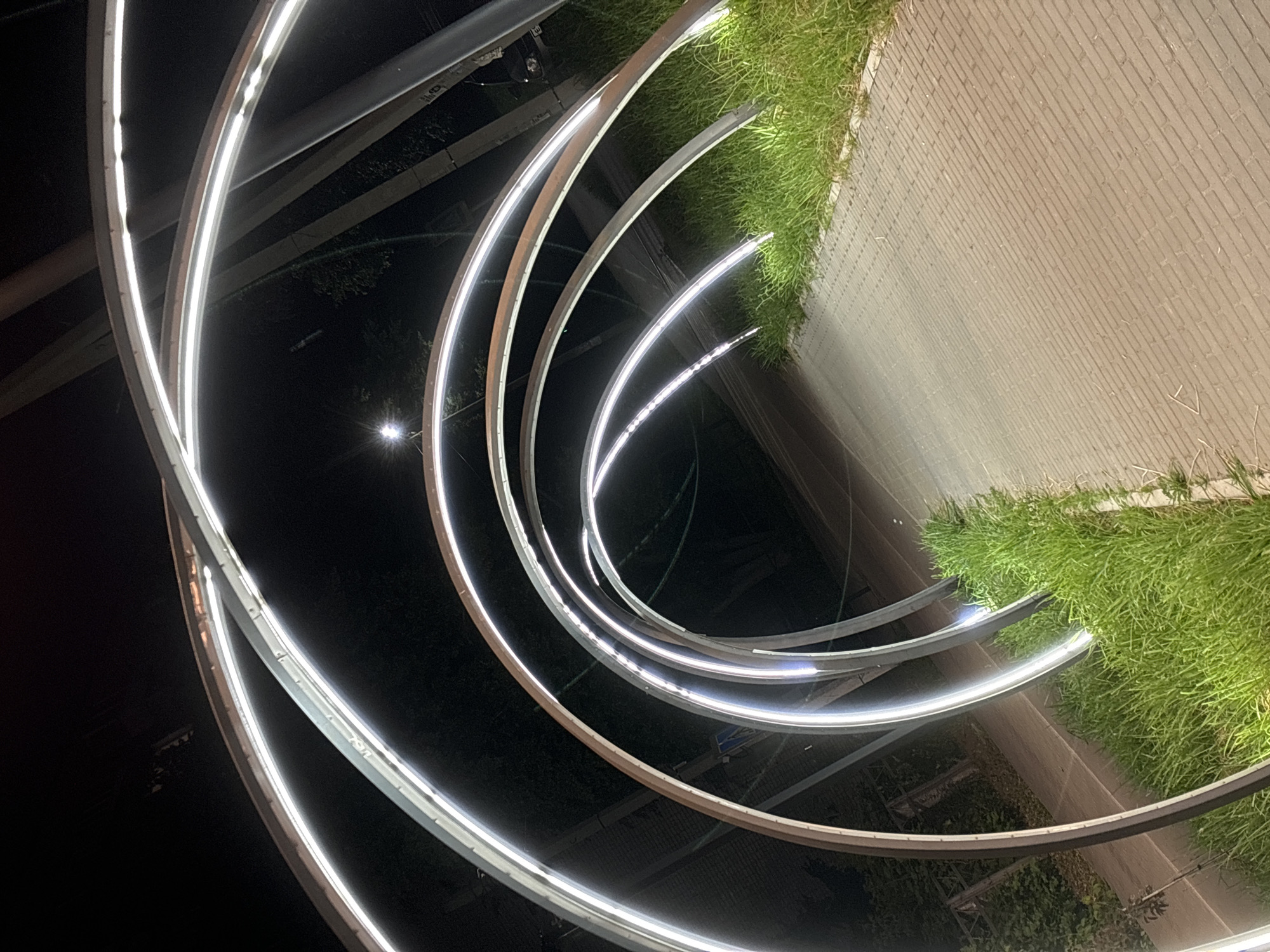 This media file depicts an outdoor art installation at night in Chaikovsky, Russia. The primary subjects are several curved, metallic structures illuminated by white LED lights. These structures are arranged in a swirling, concentric pattern, creating a dynamic visual effect. The lights are bright and modern, highlighting the sleek lines of the sculptures.
Adjacent to the art installation, there is a section of manicured green grass and a paved pathway made of light-colored bricks. The pathway is laid in a herringbone pattern. The contrast between the dark night sky, the illuminated sculptures, the vibrant grass, and the textured pathway provides a rich visual experience.
The scene is set at night, as indicated by the dark background and the artificial lighting. There are no people visible in the frame. The weather appears to be clear, allowing the artificial lights to stand out effectively. The location cues, such as the grass and brick pathway, suggest a public space like a park, plaza, or garden. There is no visible text in the image. The overall mood is modern and artistic, with a sense of quiet and calm due to the absence of activity and the nighttime setting.