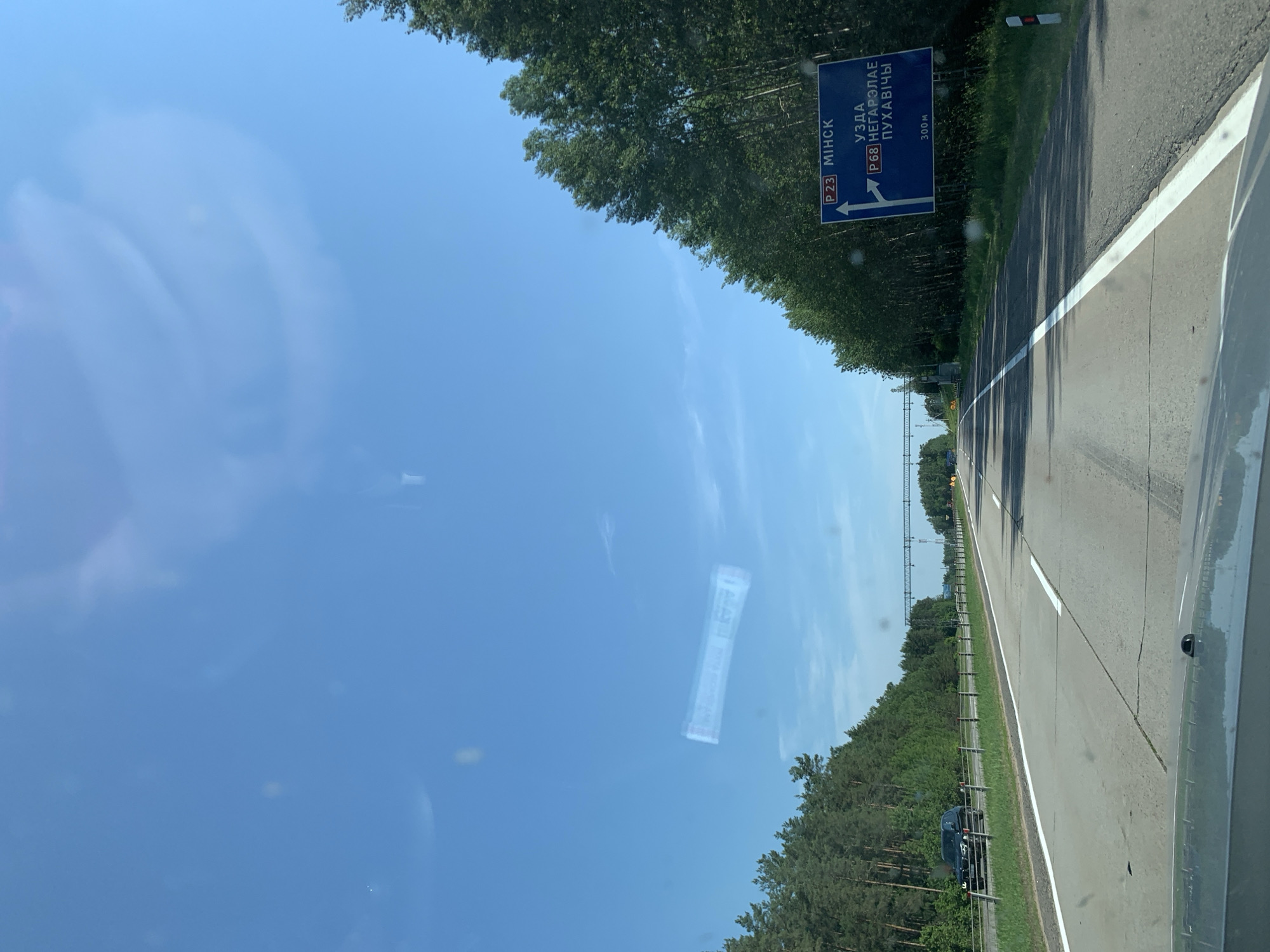 The image depicts a view from a vehicle traveling on a highway in a rural area. The sky is clear blue with some wispy clouds. On the right side of the road, a blue highway sign indicates directions. The top part of the sign displays "P23 МІНСК" with an arrow pointing straight ahead, signifying Minsk as a destination via route P23. Below this, it shows "УЗДА" and "P68 НЕГАРЭЛАЕ ПУХАВІЧЫ" with an arrow pointing to the left, indicating Uzda and directions to Negarelaya and Pukhavichy via route P68. At the bottom of the sign, "300M" is visible. The highway has multiple lanes, and trees line both sides of the road. A dark SUV is visible in the distance on the left lane. The overall scene suggests a sunny day on a road trip in Belarus, specifically within the vicinity of Гацукоўскі сельскі Савет.