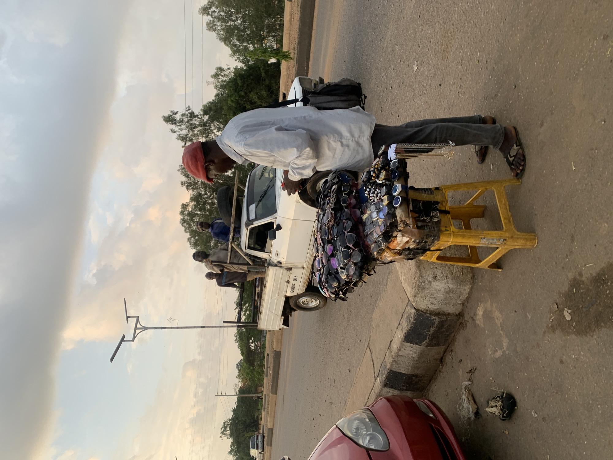 In Gura Top, Nigeria, a man wearing a red cap, white shirt, and grey trousers stands on a road next to a yellow plastic step stool laden with sunglasses and watches. He appears to be interacting with the back of a white truck; passengers are sitting in the truck's bed. The scene is captured during daylight, and a red car is partially visible in the foreground. A street lamp is visible, as are overcast skies and trees in the background.