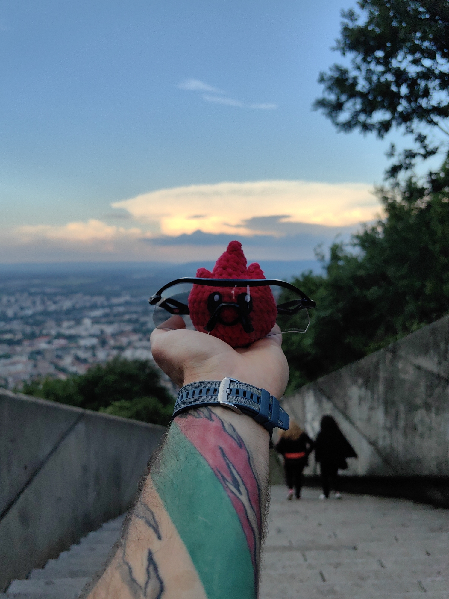 A close-up shot features a hand holding a red crocheted figure with sunglasses, superimposed against a cityscape, which is Shumen, Bulgaria, seen from the Monument to 1300 Years of Bulgaria memorial complex. The viewpoint is from an elevated position on the monument complex, looking out over the city. The sky is visible in the background, displaying a mix of blue hues and sunset colors. A wristwatch is on the person's arm, which also shows tattoos. In the background, two figures are walking down a staircase. Concrete retaining walls surround the walking path.