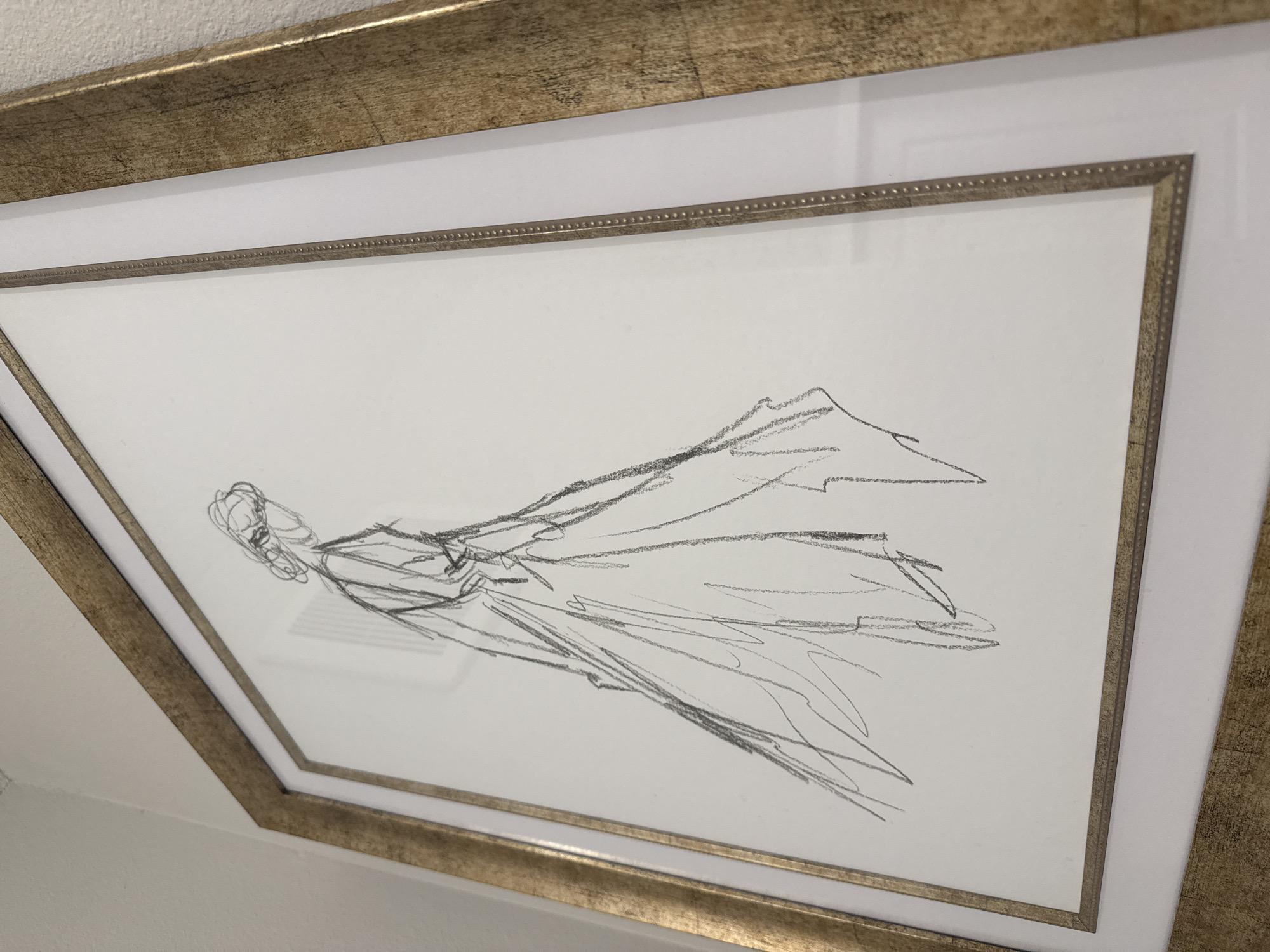 A framed charcoal or graphite drawing hangs on a white wall. The frame consists of two layers: an outer, wider frame in gold leaf, and an inner, thinner frame in silver or light gold with a beaded edge. The drawing depicts a figure in motion. The figure appears to be wearing flowing clothing. The drawing is an expressive sketch with loose lines. The scene is indoors. The location is likely in Orlando, United States, given the prompt's instructions.