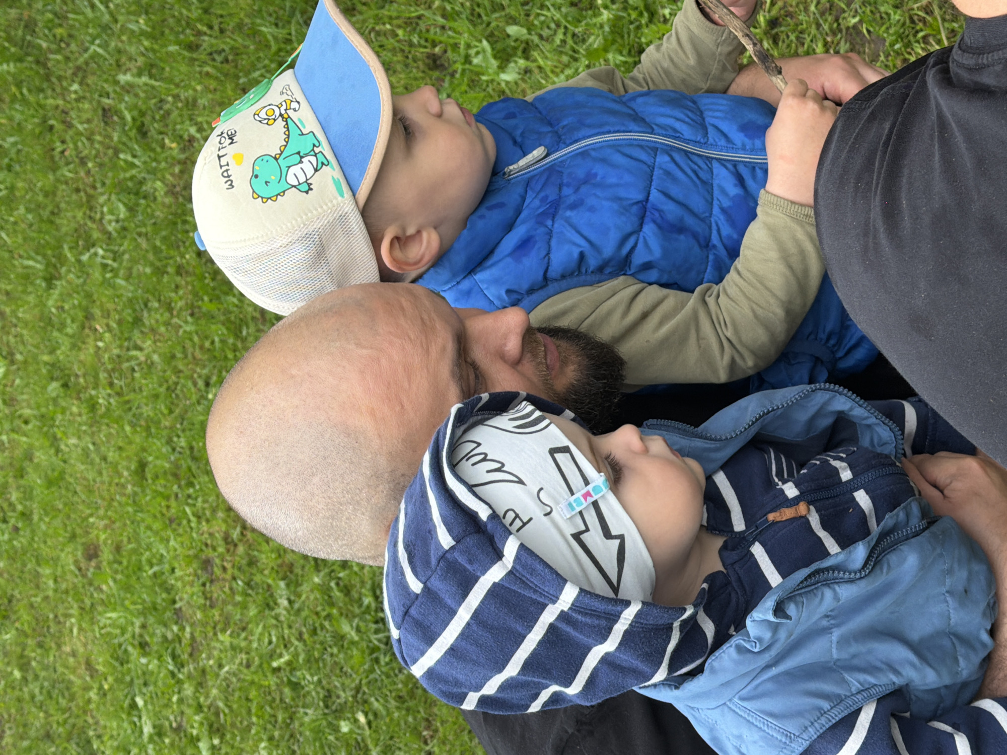 The image depicts a close-up, medium shot of a man and two young children outdoors, likely in Votkinsk, Russia. The background is predominantly green grass, suggesting a park or grassy area.
In the foreground, a man with a shaved head and a beard is holding a child. The man is wearing a dark shirt. The child he is holding is wearing a blue hooded jacket with white stripes, and a light-colored beanie with black lightning bolt graphics. The child's face is partially obscured, but their left eye is visible.
To the right of the man, another child is seated, wearing a blue puffer vest over a long-sleeved shirt and a beige baseball cap with a cartoon dinosaur graphic and the text "WAIT FOR ME". This child is looking to their right. The child's right hand is holding a small stick.
The overall impression is one of a casual outdoor scene with a family.