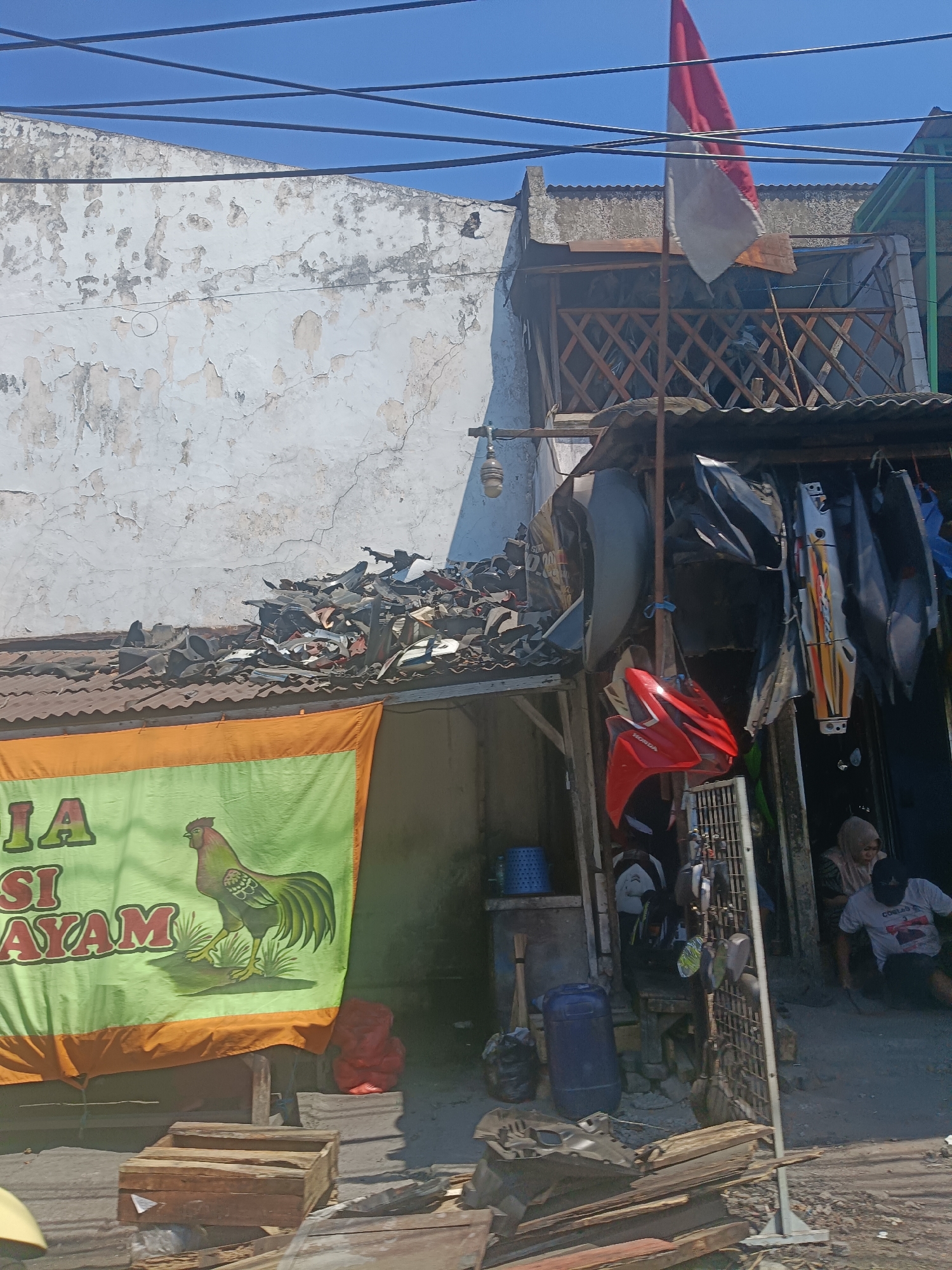 This image depicts a street scene in Surabaya, Indonesia, characterized by a weathered building facade and an assortment of items displayed outside what appears to be a shop. The building has a white, peeling plaster exterior under a clear blue sky, with electrical wires strung across the top.
In the foreground, a large green banner with orange trim is visible. The banner features text and a graphic of a rooster. To the right of the banner, a collection of motorcycle parts is displayed on a roof and hanging from a makeshift structure. These parts include various fairings, fenders, and other components in different colors, primarily gray, black, white, and red. One prominent red motorcycle fairing hangs prominently.
Behind the motorcycle parts, the entrance to a shop is visible. Two individuals are partially visible within this entrance: one person wearing a hijab and a light-colored garment, and another person wearing a dark cap and a light-colored t-shirt with writing on it. They appear to be sitting or interacting within the shop.
The ground level of the scene shows scattered debris, including pieces of wood, a blue plastic barrel, and other assorted items, suggesting a working or storage area. The overall impression is of a busy, possibly informal commercial area within the city.