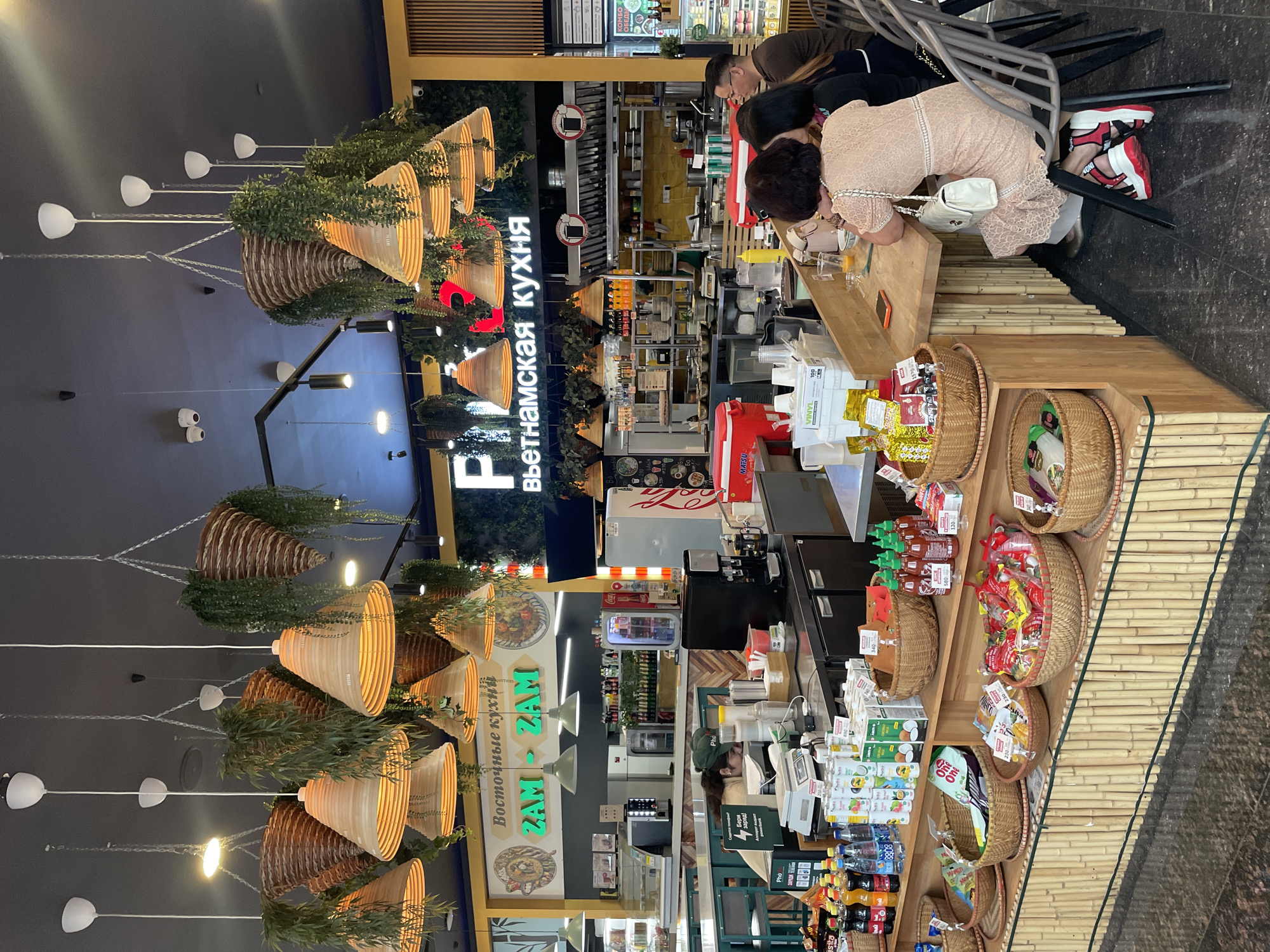 The image shows the interior of a Vietnamese restaurant. In the foreground, there are several woven baskets filled with snacks and drinks. A counter displays various food items and beverages for sale. Behind the counter, a woman wearing a green cap is working at a register. In the background, a man and two women are seated at the counter, engaged in conversation. The restaurant features decorative hanging plants and woven light fixtures. The overall lighting is warm and inviting, with a focus on natural materials like bamboo and wood.