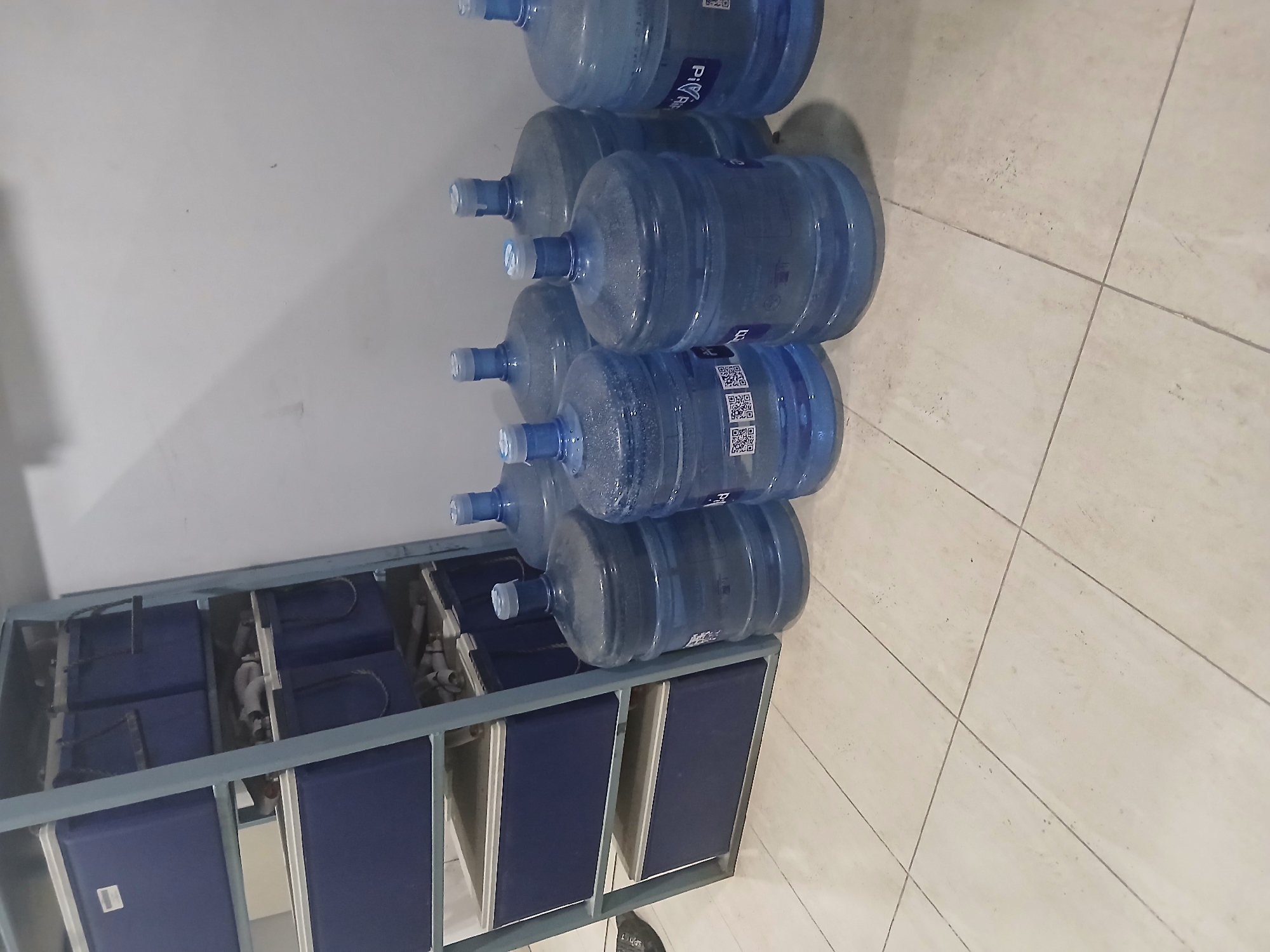 The image shows a storage area with a metal shelving unit on the left, filled with blue containers. On the right, several large, blue plastic water jugs are arranged on a tiled floor against a white wall. The jugs are opaque and appear to be full or partially full. Some of the jugs have labels with QR codes. The overall environment suggests a storage or stockroom, potentially in Peshawar City Tehsil, Pakistan.