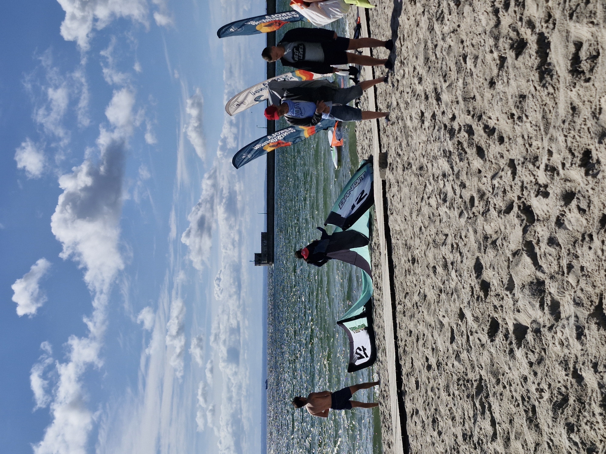 The image shows a sunny day at a beach in Hel, Poland, with the Baltic Sea in the background. Several people are present on the sandy shore, some of whom are involved with kitesurfing equipment. In the foreground, there are several kites laid out on the beach. Two men are standing near a large kite, one wearing a red cap and a blue t-shirt, and the other wearing a dark jacket and shorts. To the left, a young man is standing in the shallow water, facing the sea. Further out in the water, there is a structure that appears to be a pier or a lighthouse, and a small boat. The sky is blue with scattered white clouds. The scene suggests a recreational setting related to water sports.