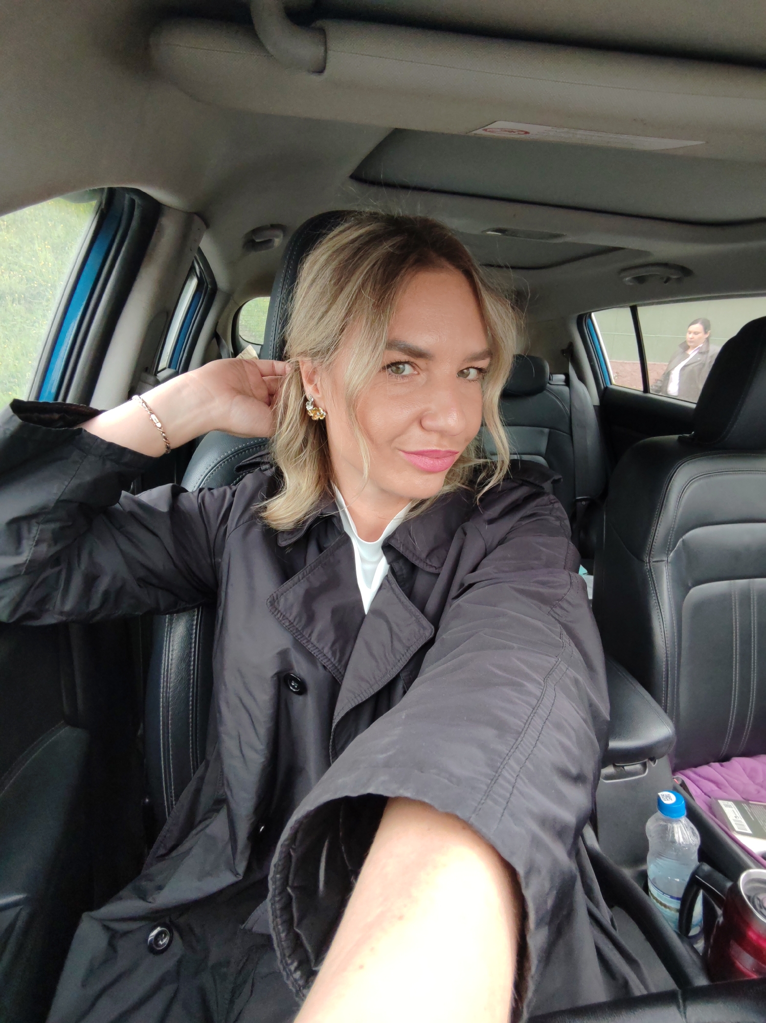 A medium shot captures a blonde woman with fair skin, green eyes, and light pink lipstick, posing for a selfie inside a car. She is wearing a black trench coat over a white shirt and a bracelet on her left wrist. She has a gold earring with a floral design in her right ear. The car's interior is visible, with black leather seats and a sunroof. In the background, a woman with dark hair wearing a dark jacket over a white shirt is seen through the car window. The environment outside the car appears to be an overcast day with greenery visible. A water bottle and a red thermos are present in the car's center console.