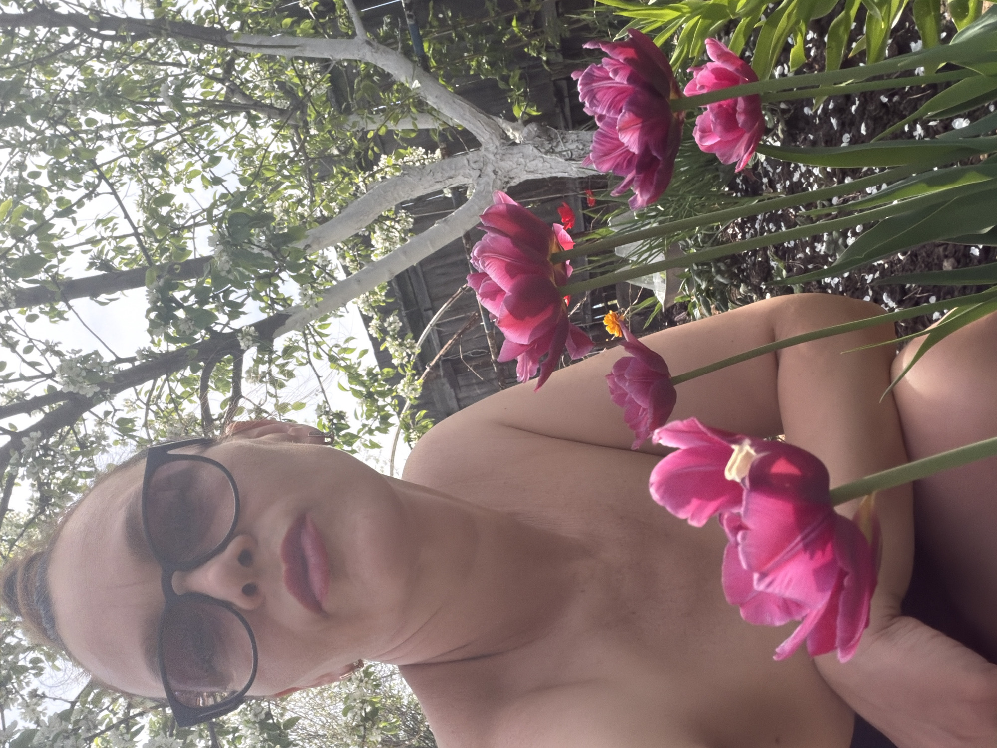 A person wearing sunglasses is facing the camera. Pink tulips are in the foreground and a tree with white branches and green leaves is in the background. The location is Chaikovsky, Russia.