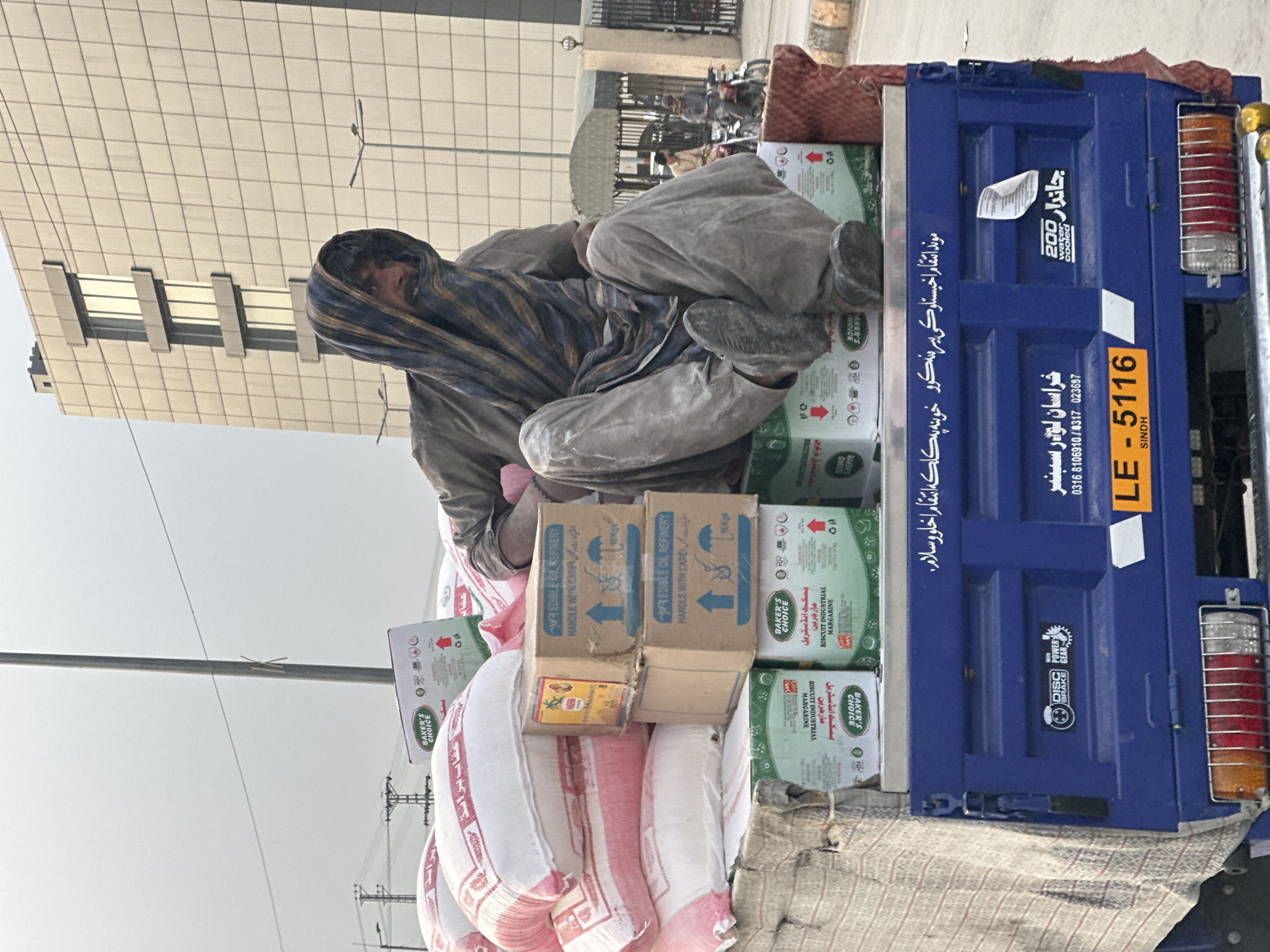 A man with a patterned scarf draped over his head sits atop a load of goods on the back of a blue truck. He is wearing dusty, neutral-colored clothing. The goods consist of sacks of white material with red stripes and several boxes labeled "BAKER'S CHOICE BISCUIT INDUSTRIAL MARGARINE" and "WR EDIBLE OIL REFINERY HANDLE WITH CARE". The truck, partially visible, has the license plate "LE-5116 SINDH" and various Urdu writings. In the background, a modern building with a grid-like facade and a street scene with motorbikes are visible. The photo appears to have been taken during the day in an urban setting. Considering the information, this image was likely captured in Pakistan (SINDH) on the back of a blue truck. The city is not Quetta but a city located in Sindh, Pakistan.
