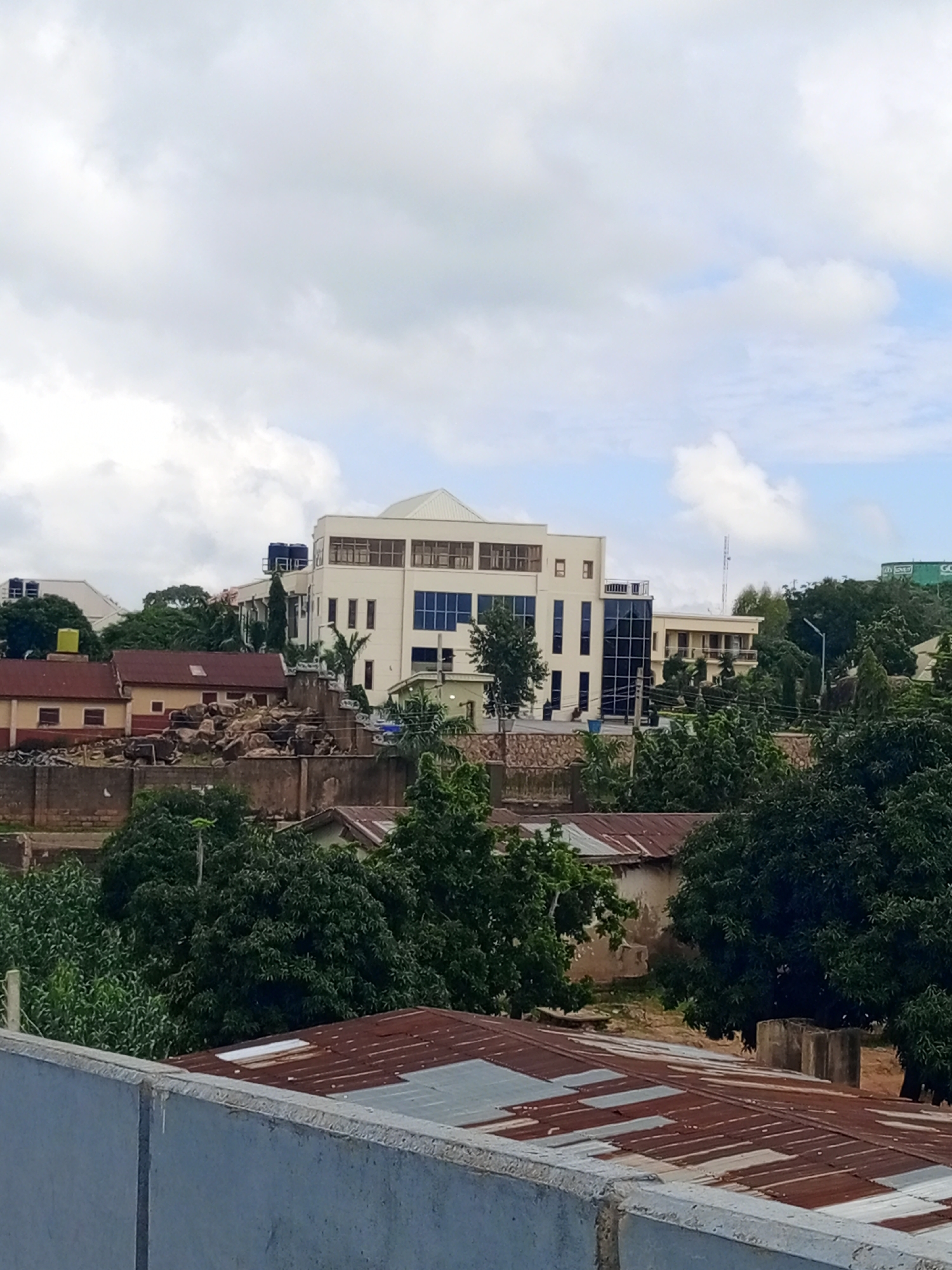 The image captures an overhead view of a cityscape in Jos, Nigeria, dominated by buildings, trees, and a cloudy sky. A large, cream-colored building with numerous windows, some tinted blue, stands prominently in the center, featuring a modern architectural style with multiple levels and a sloped roof. Adjacent to it are smaller, older buildings with red-tiled roofs and yellowish walls. A stone wall runs across the mid-ground, separating different property levels. Dense green foliage from various trees and shrubs is scattered throughout the scene, indicating a vegetated urban environment. In the foreground, a rusty, corrugated metal roof is visible, suggesting older or less maintained structures. The overall impression is of a developing urban area with a mix of modern and older constructions, set against a backdrop of natural greenery under an overcast sky.