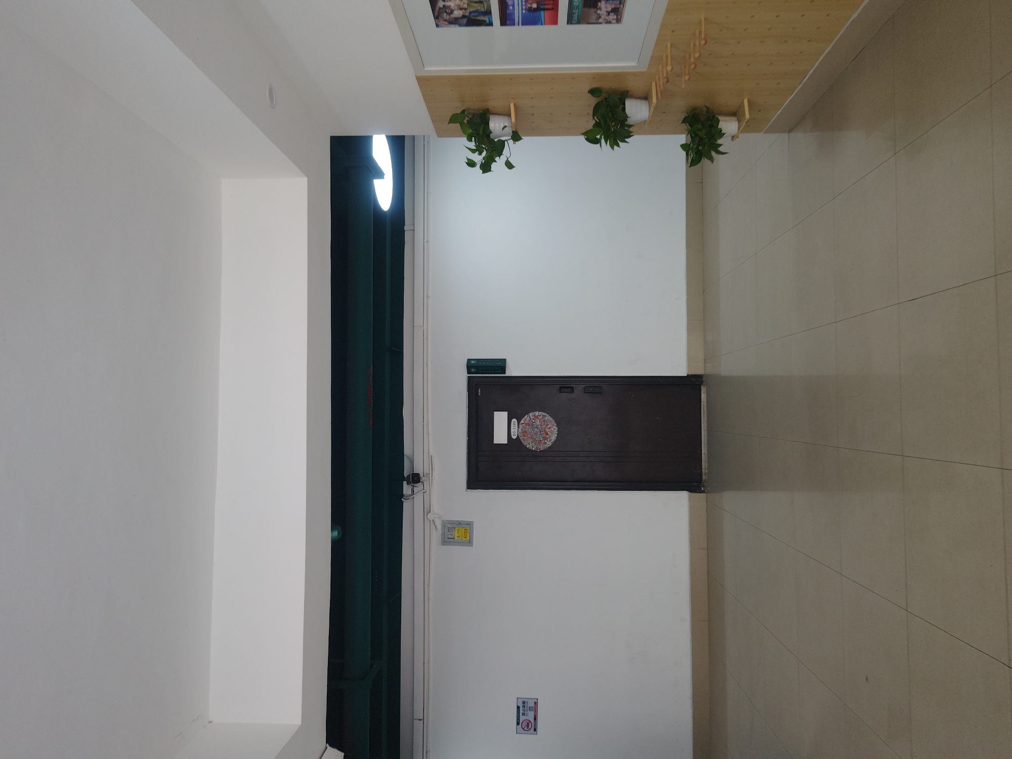 The image shows an indoor hallway with a tiled floor. On the left side of the hallway, there is a white wall with a "no smoking" sign and an electrical panel. In the center of the hallway, there is a dark brown door with a circular decorative plaque. To the right of the door, a white framed bulletin board is mounted on a light brown wooden paneling. Several small potted plants are displayed on wooden shelves attached to the bulletin board. The ceiling is white and has a lighting fixture visible. The overall environment appears to be a clean and well-maintained indoor space. The image is not sufficient to determine the specific location within the Chongqing High-tech Zone, China.