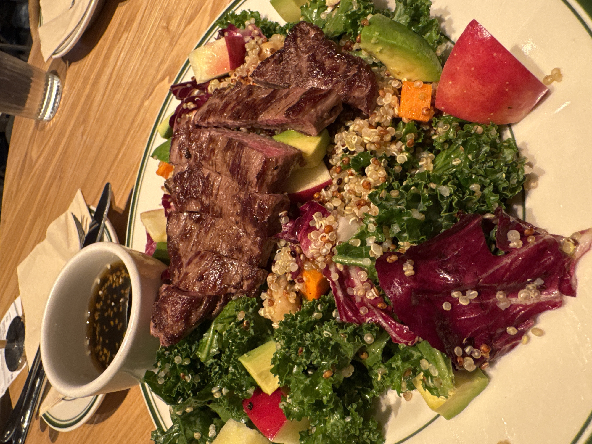 The image shows a plate of food on a wooden table. The main component is sliced steak, which is placed on top of a salad. The salad consists of kale, red lettuce, quinoa, avocado, apple slices, and diced carrots. A small white cup containing a dark sauce with sesame seeds sits to the left of the plate. There is a glass of water, a fork, a knife, and a napkin visible as well. The plate is white with a green rim. The location is not evident from the image, so providing a precise location like Yuzhong District, China, is not possible.