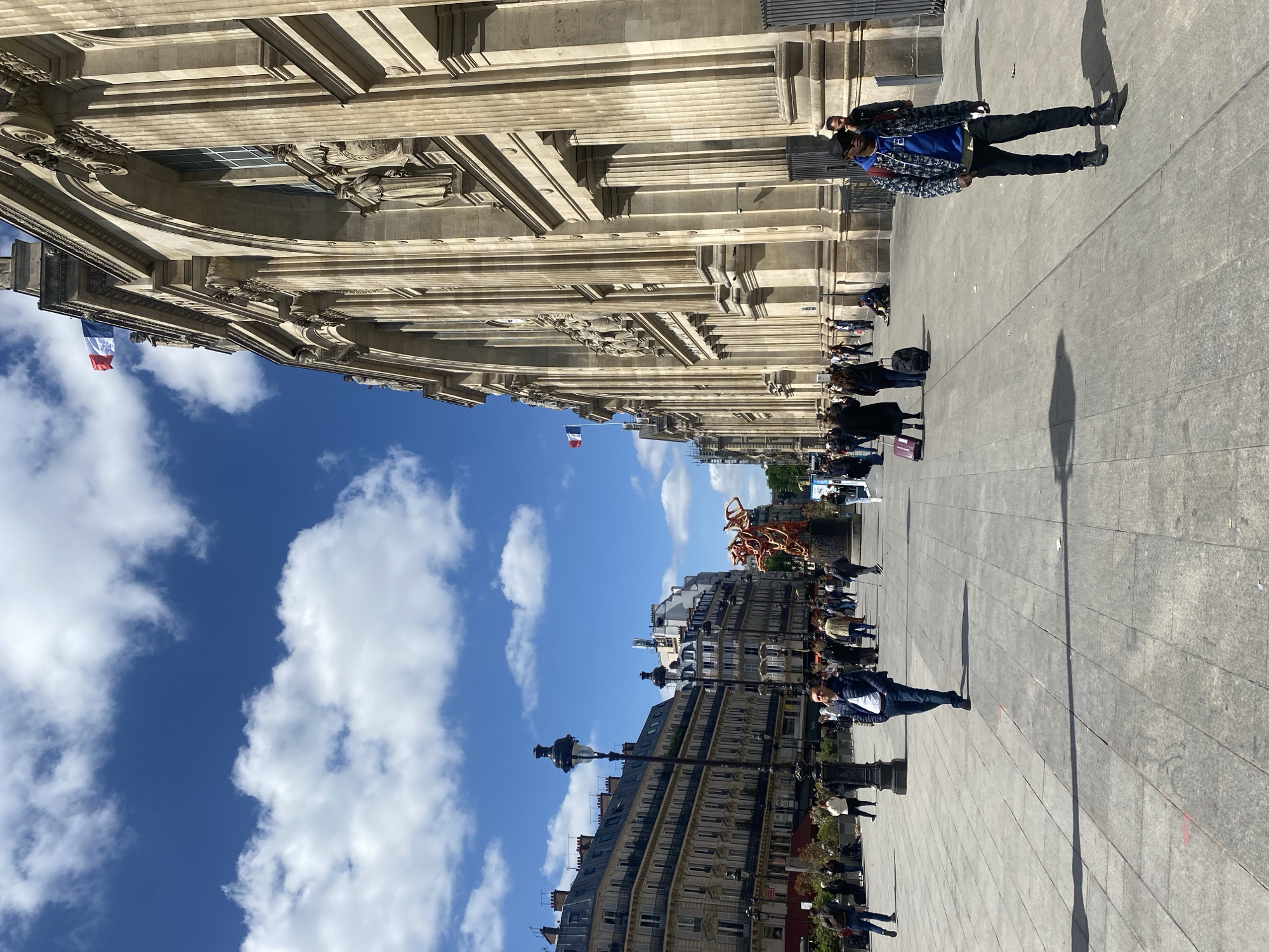 This image captures a vibrant street scene in Paris, France, on a clear, sunny day. The perspective looks down a wide, paved pedestrian area.
On the left, a grand, classical-style building dominates the frame, featuring elaborate stone facades, numerous columns, and decorative sculptures. Several French tricolor flags are visible, fluttering from poles attached to this impressive structure, serving as a clear geographical cue.
Across the pedestrian expanse, on the right side, stands a row of characteristic Parisian apartment buildings with multiple stories, dark roofs, and numerous windows. A distinctive, abstract sculpture, predominantly orange and red, is visible in the middle distance, adding a modern artistic touch to the historic surroundings.
The pedestrian area is bustling with people. Individuals and small groups are seen walking in various directions, some carrying luggage, suggesting tourism or daily commutes. Two men are prominent in the foreground: one in a blue hoodie and patterned jacket, and another further back in a dark jacket. Long shadows cast by people and street lamps indicate that it is either mid-morning or early afternoon. The sky above is a brilliant blue with scattered white cumulus clouds, contributing to the bright and inviting atmosphere. No discernible text is visible on any buildings or signs.