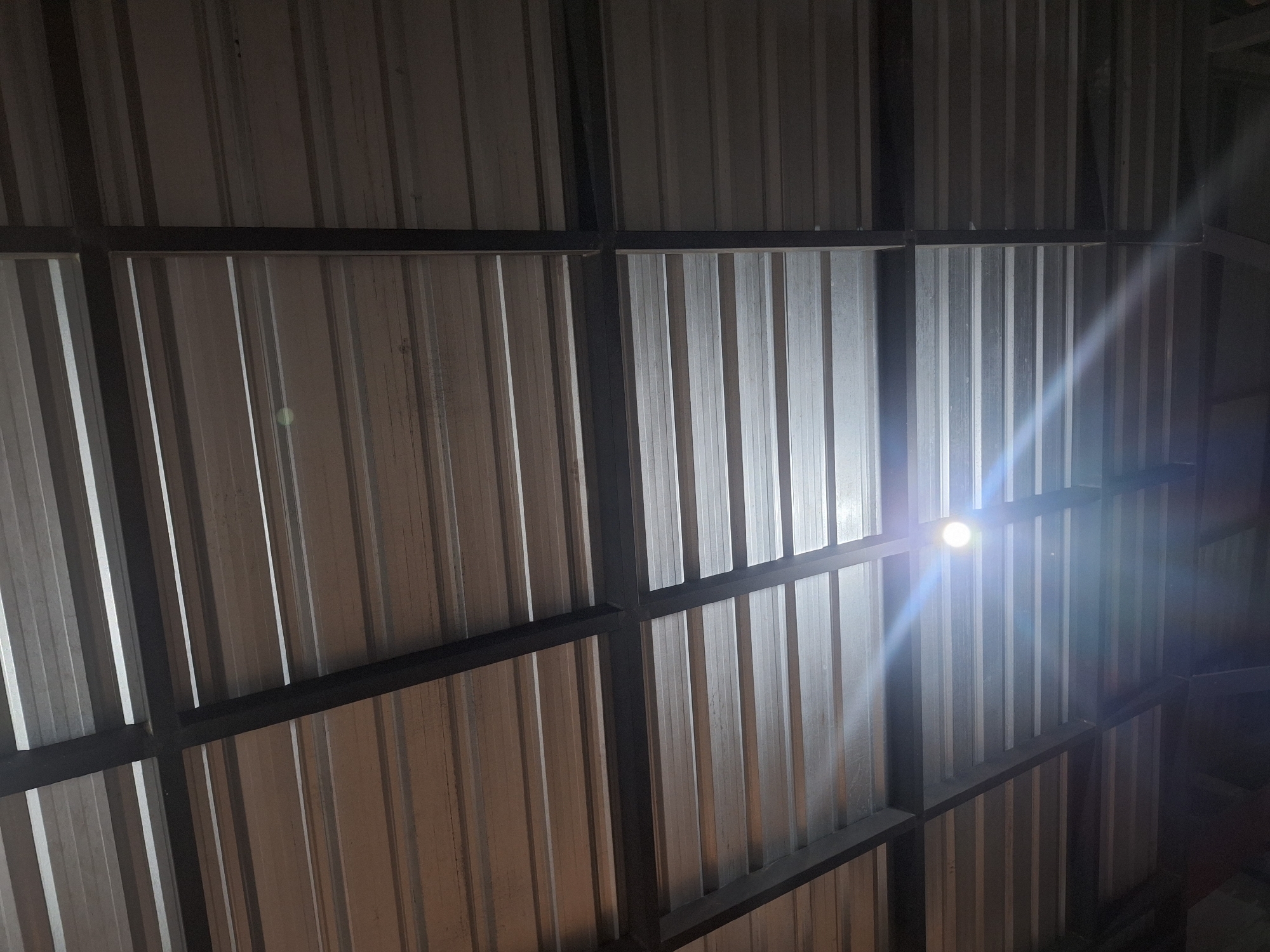 The image shows a corrugated metal roof with a dark metal frame. The roof panels are oriented horizontally, and the frame creates a grid pattern. A bright light source is visible in the lower left quadrant of the image, causing a lens flare and illuminating the surrounding metal panels. The environment appears to be indoors or under some kind of shelter, given the roofing material and lighting. The scene is located in Samarinda, Indonesia, and depicts the interior of a structure with a visible roof. There are no people or discernible actions taking place in the image.