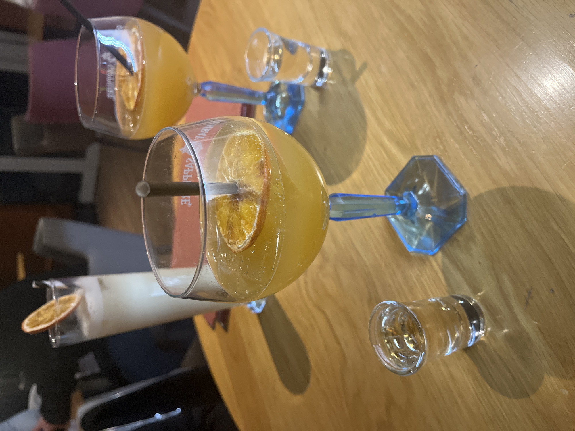 The image depicts a tabletop scene, likely in a bar or restaurant. There are three cocktails in stemmed glasses, each garnished with a slice of citrus, most likely orange. The stems of the glasses are blue. One cocktail appears to be a lighter color, possibly a creamy concoction, while the other two are a vibrant orange hue. Two small shot glasses, each containing a clear liquid, are also present on the table. The tabletop is a light, natural wood grain, suggesting a casual setting. The location is Montrond-les-Bains, France. There are no people visible in the immediate frame, but hints of chairs and clothing suggest the presence of patrons. The environment is well-lit, highlighting the textures and colors of the drinks and the table surface.