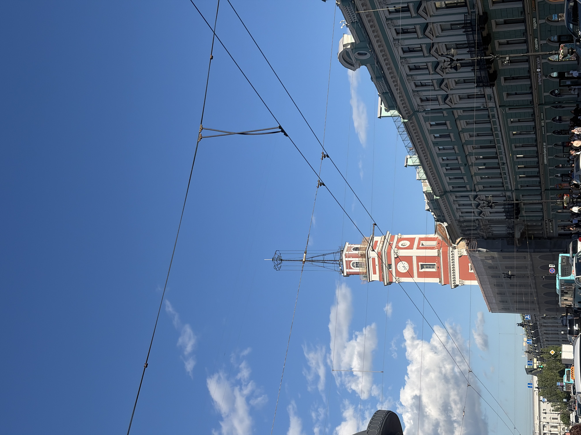 This is a photograph taken from a street-level perspective in Saint Petersburg, Russia. The image is dominated by a vast expanse of blue sky with scattered white clouds. Several overhead wires, likely for public transportation, crisscross the frame, creating a geometric pattern against the sky.
In the mid-ground, a prominent red and white clock tower stands tall. The tower has architectural details, including arched windows and a clock face. The building appears to be part of a larger complex. Adjacent to the tower, on the right side of the image, is a multi-story building with a green facade, characterized by its classical architectural style, featuring numerous windows and decorative elements. This building lines the street, suggesting an urban setting.
On the street level, several vehicles are visible, including a blue bus and other cars, indicating ongoing traffic. People can be seen on the sidewalks, though they are small in the frame. The presence of street signs and traffic lights further confirms the urban environment.
The bright sky and clear visibility suggest that the photograph was taken on a sunny day, likely during daylight hours. The style of architecture and the general urban landscape are consistent with the historical city of Saint Petersburg. The overall mood of the image is one of a vibrant, active city on a pleasant day. Some text is visible on the green building, though it is difficult to read clearly due to the image resolution and orientation.