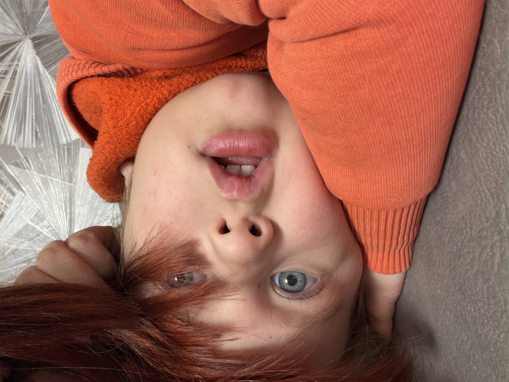 A close-up, rotated, portrait of a child with light skin, blue eyes, and red hair. The child is wearing an orange hooded sweater. The background is a mottled gray surface. The location is Chaikovsky, Russia.
