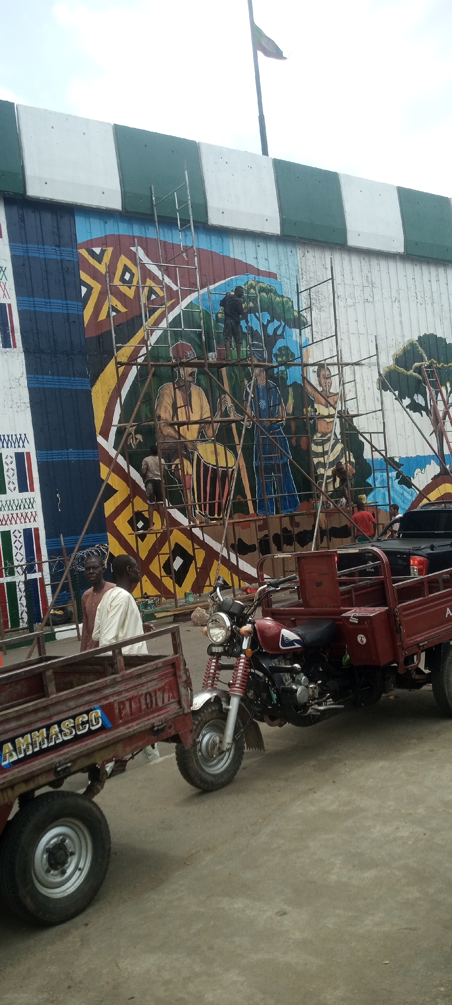 The image depicts a street scene in Jalingo, Nigeria, in front of a large mural. The mural, which appears to be under construction or being painted, features a colorful design with geometric patterns and depictions of people engaged in activities like drumming. Scaffolding is erected in front of the mural, with several individuals working on it.
In the foreground, a red motorized three-wheeled vehicle, commonly known as a "keke," is parked, with its cargo bed empty. Another keke, slightly to the left, also appears to be empty. A motorcycle, also red, is parked next to the first keke. Two men are standing near the first keke, observing the mural. One man is wearing a brown traditional outfit, and the other is in a white outfit. A black pickup truck is partially visible behind the red keke and motorcycle.
The environment appears to be an outdoor public space, possibly a street or market area. The sky is overcast. The overall impression is one of ongoing activity, with the mural painting being a central focus. The presence of the kekes suggests a transportation hub or a commercial area.