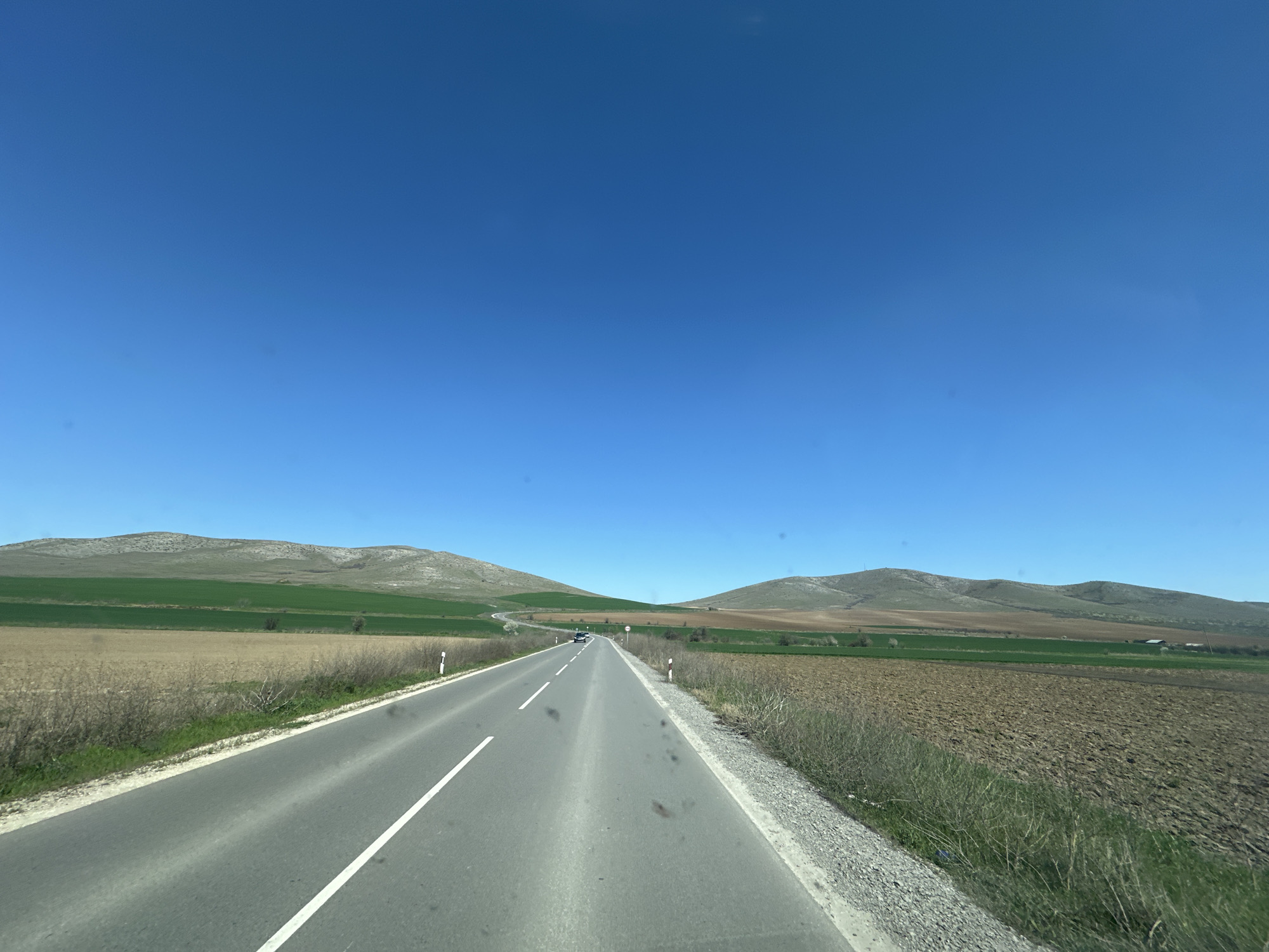 The image depicts a straight, two-lane asphalt road leading into the distance. A white dashed line divides the lanes. The sky is a clear, cloudless blue. On either side of the road, there are fields, some brown and possibly plowed, and others green. Low, rolling hills are visible in the background. The road appears to be somewhere near Isperihovo, Bulgaria.