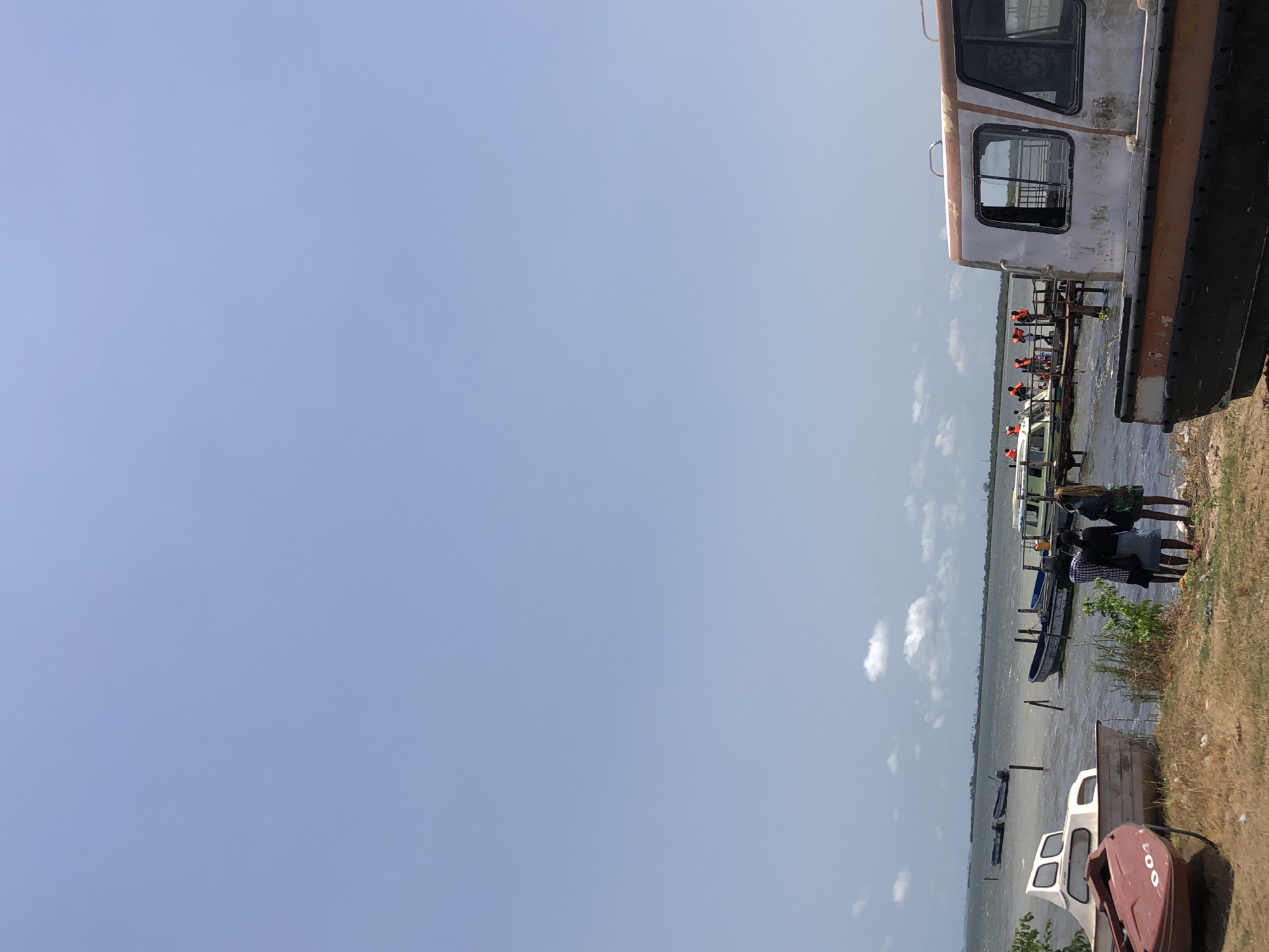 The image shows a waterfront scene. A large, rusty boat sits on the shore to the right, its windows visible. People wearing orange life vests sit in a row on a dock or smaller boat. Other boats are docked or in the water. People stand along the shore, near some grass and debris. The sky is blue with a few scattered clouds. The location is near a body of water, with the city listed as Unknown, Unknown.