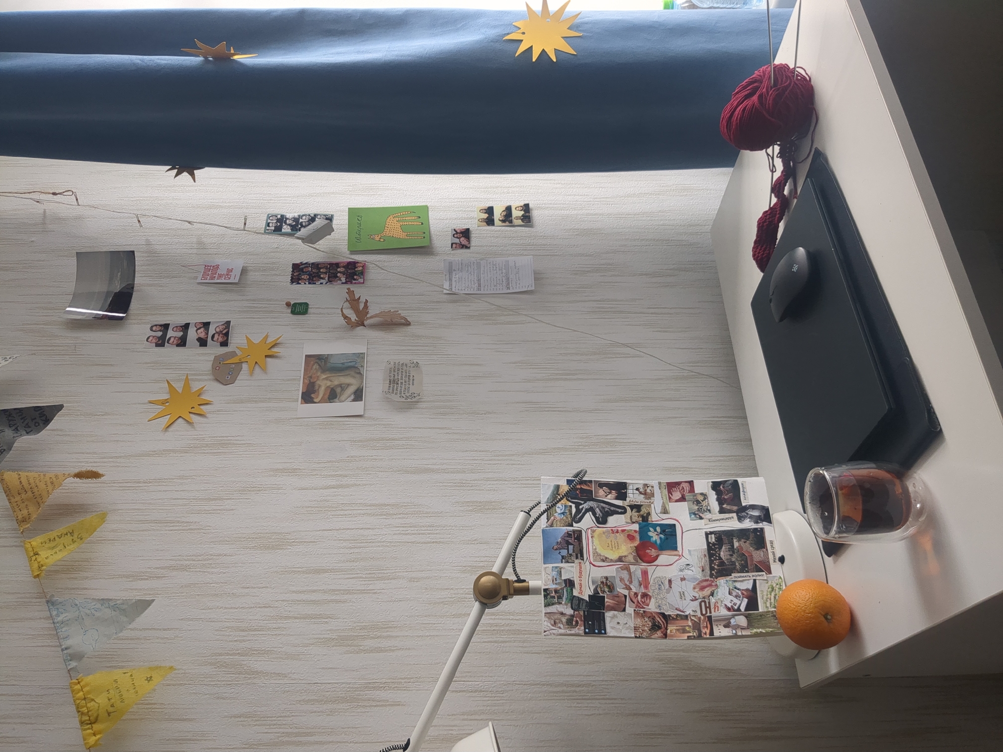 A slightly angled high-angle shot captures a room or workspace, seemingly in Saint Petersburg, Russia, based on the Cyrillic text visible on some of the displayed items. A dark blue curtain or fabric is draped across the top, adorned with paper star cutouts. Below the curtain, a white wall serves as a makeshift display area. A string of fairy lights runs horizontally across the wall. Various items are pinned or taped to the wall, including photographs, postcards, handwritten notes, and small decorative elements, such as paper leaves and stars. A pennant banner hangs on the left side.
To the right, a white desk surface is visible. On the desk is a black laptop or tablet, a gray computer mouse, a ball of red yarn, a glass containing a dark liquid, and a single orange. A white desk lamp is positioned near the center. The lamp's shade is also covered with a collage of images and text. The overall impression is of a creative and personalized space.