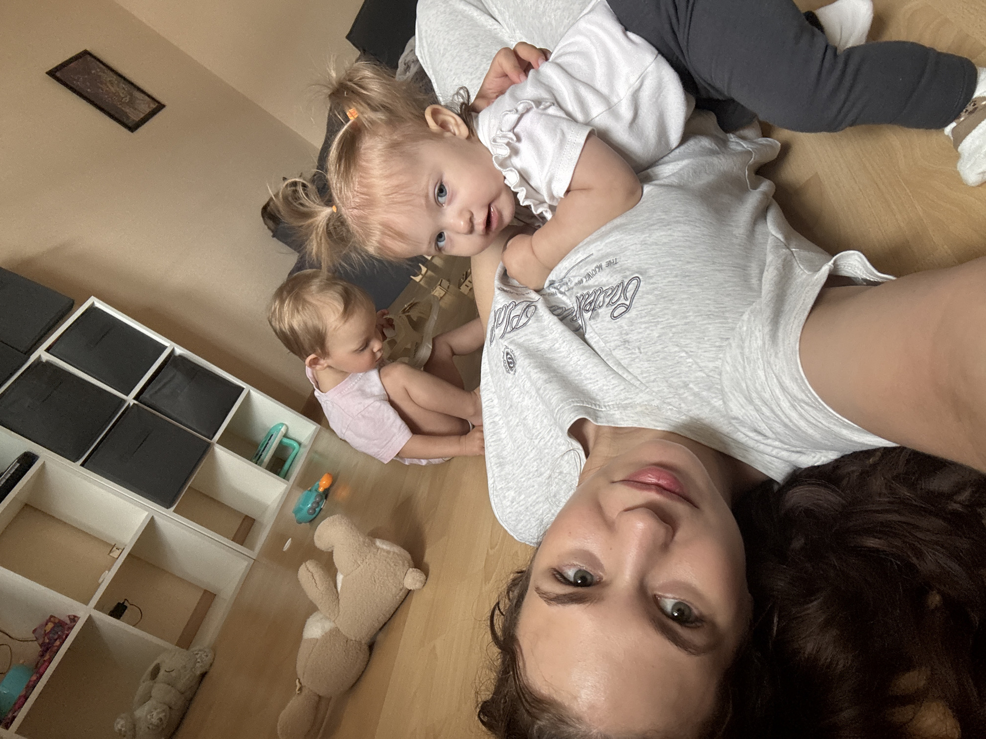 An indoor, eye-level, medium shot captures a woman and two young children on a wooden floor. The woman, positioned in the foreground, is lying on her back with her head resting on her dark hair. She looks up at the camera with a slight smile. She is wearing a gray t-shirt with a graphic design. Partially visible is a toddler wearing a white ruffled shirt and gray pants, leaning against the woman and looking toward the camera. Another child, also in a pink t-shirt, is further in the background, crouched down and facing away from the camera. In the background, a white shelving unit with black storage bins is visible, along with a stuffed animal and a toy. The lighting is soft, suggesting ambient room light. The overall scene appears to be a domestic environment.