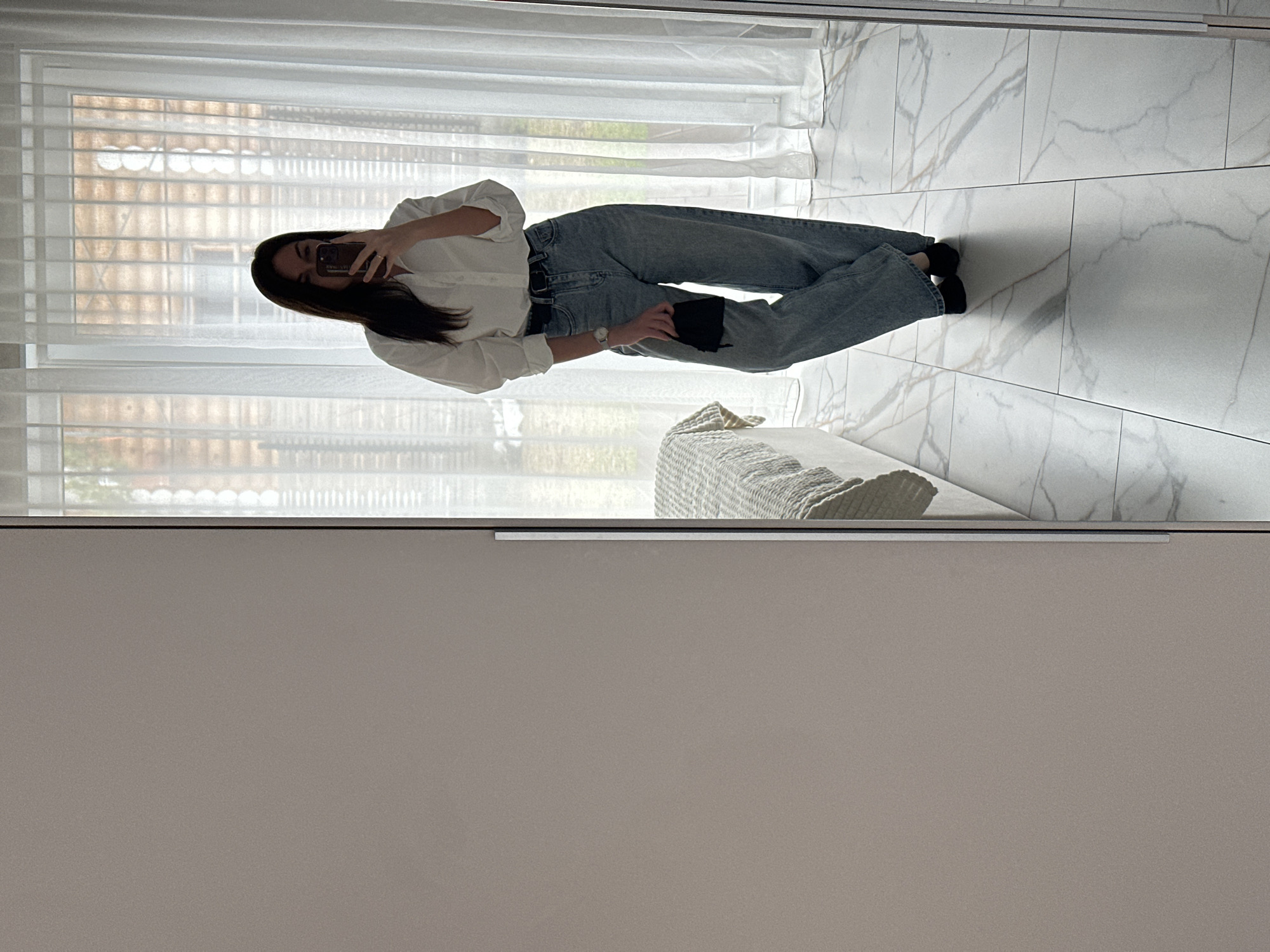 A young woman is captured in a mirror selfie, standing upright in what appears to be a modern indoor setting in Chaikovsky, Russia. She has long, dark hair and is dressed in a white long-sleeved button-up shirt, light-wash blue high-waisted jeans cinched with a dark belt, and black socks. Her right hand holds a dark-colored smartphone, positioned to take the photo, partially obscuring her face. On her left wrist, she wears a watch with a metallic strap, and in her left hand, she holds a small, rectangular black object, possibly a wallet or clutch.
The background reveals a bright room, illuminated by natural light entering through a large window covered by sheer white curtains and horizontal blinds. Beyond the window, a blurred outdoor view suggests buildings or foliage. The floor is covered with large rectangular white tiles featuring prominent grey and light brown marble-like veining. To the right, a textured, light-colored throw blanket or cushion rests on what appears to be a piece of furniture, possibly a bed or sofa. The lower portion of the image shows a flat, light grey-beige surface with a sleek metallic handle, indicating the reflective surface is part of a large wardrobe or cabinet. The overall ambiance is clean and minimalist, suggesting a contemporary living space. No discernible text or explicit emotions are visible.