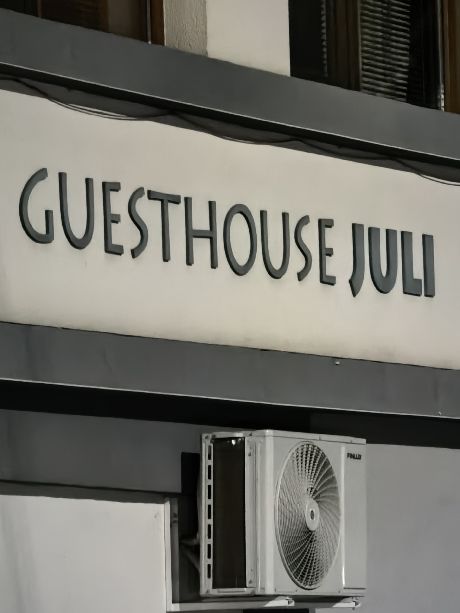 The image shows a sign reading "Guesthouse Juli" in a dark gray, sans-serif font. Below the sign is a white Finlux air conditioning unit. The location is unspecified.
