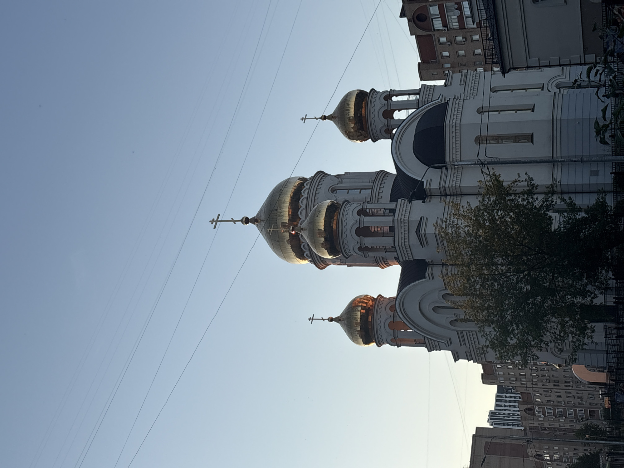 The image shows a view of an Orthodox church with golden domes against a clear blue sky. The church has white walls and intricate architectural details. Several domes are visible, each topped with a cross. The sun is shining, casting a warm glow on the domes, suggesting it is either morning or late afternoon. The low angle of the sun also creates some shadows.
In the background, there are tall apartment buildings, indicating an urban setting. Some trees with green leaves are also visible, partially obscuring parts of the church and the apartment buildings. Several power lines stretch across the frame, crossing in front of the sky and the buildings.
The image is oriented vertically. There is no discernible text in the image. The scene appears peaceful and serene, with the sunlit domes of the church standing out against the urban landscape and the clear sky.