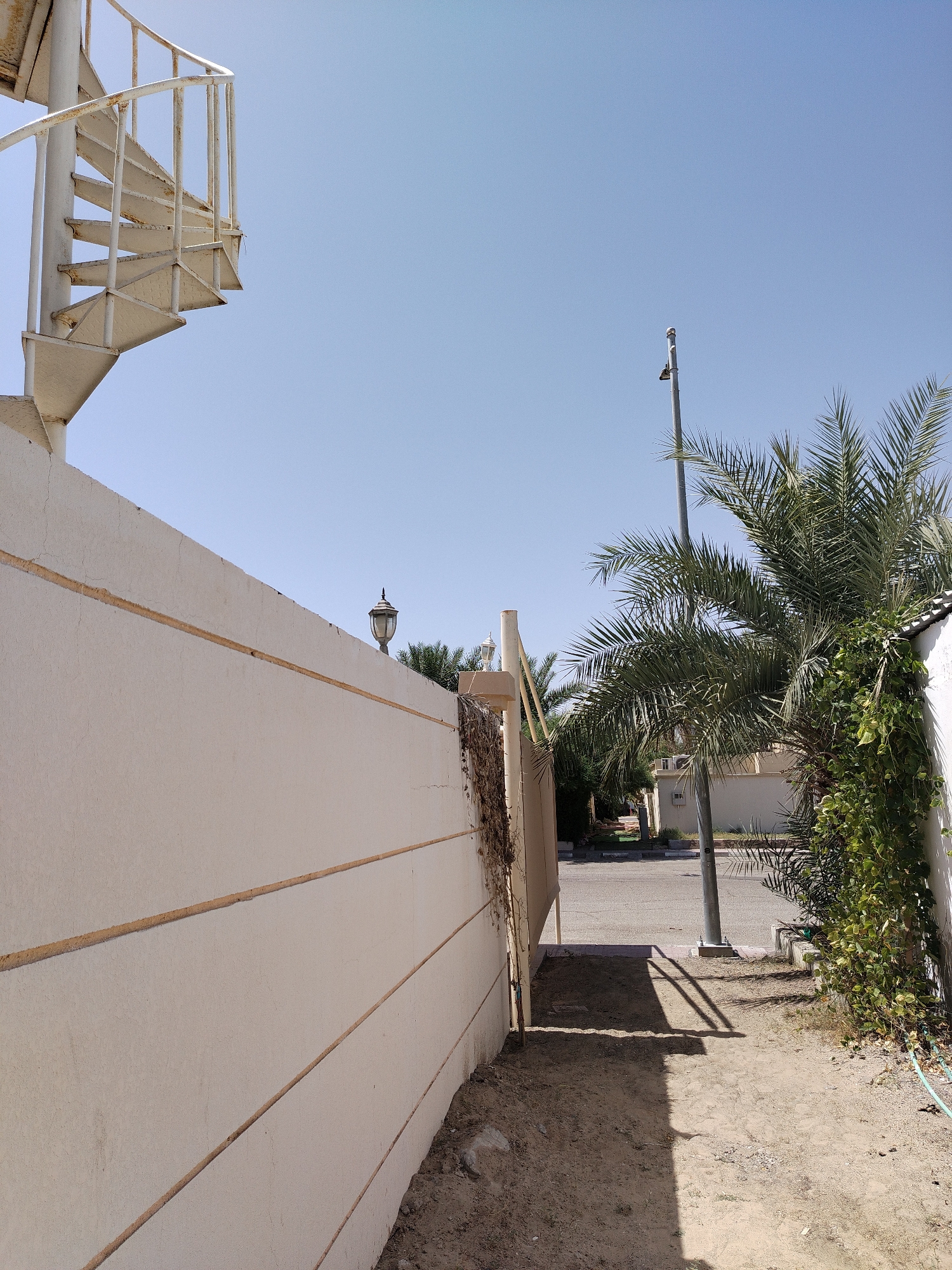 A medium shot shows an exterior view of a residential neighborhood in Al Mas'oudi, United Arab Emirates. A cream-colored wall runs along the left side of the frame. A white metal spiral staircase is partially visible at the top left of the wall. A light post is centered in the image in line with a palm tree to the right. A light-colored one-story building can be seen down the street, between the wall and the tree. The ground is dirt and sand.