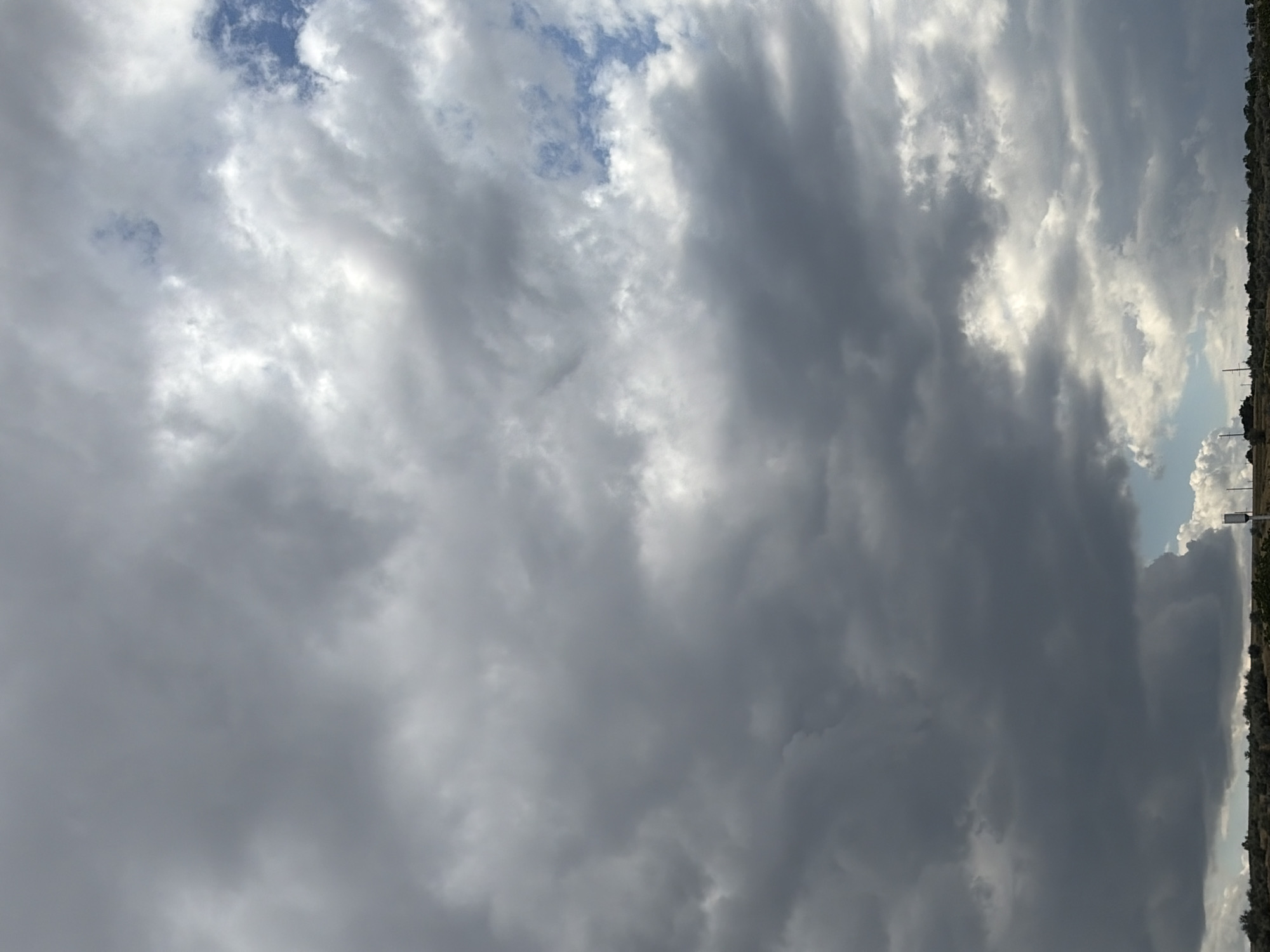 The image displays a cloudy sky above a rural landscape. The clouds are varied in shade, with some appearing dark and ominous, while others are lighter and break apart to reveal glimpses of blue sky. The foreground shows a low horizon line with sparse vegetation and some indistinct structures, possibly power lines or telecommunication towers. The scene suggests an outdoor environment, potentially in the countryside. The provided location, Oxentea, Moldova, places the image in a specific geographical context. There are no people or discernible actions or interactions within the frame. The overall impression is of a dynamic sky, possibly indicative of changing weather.
