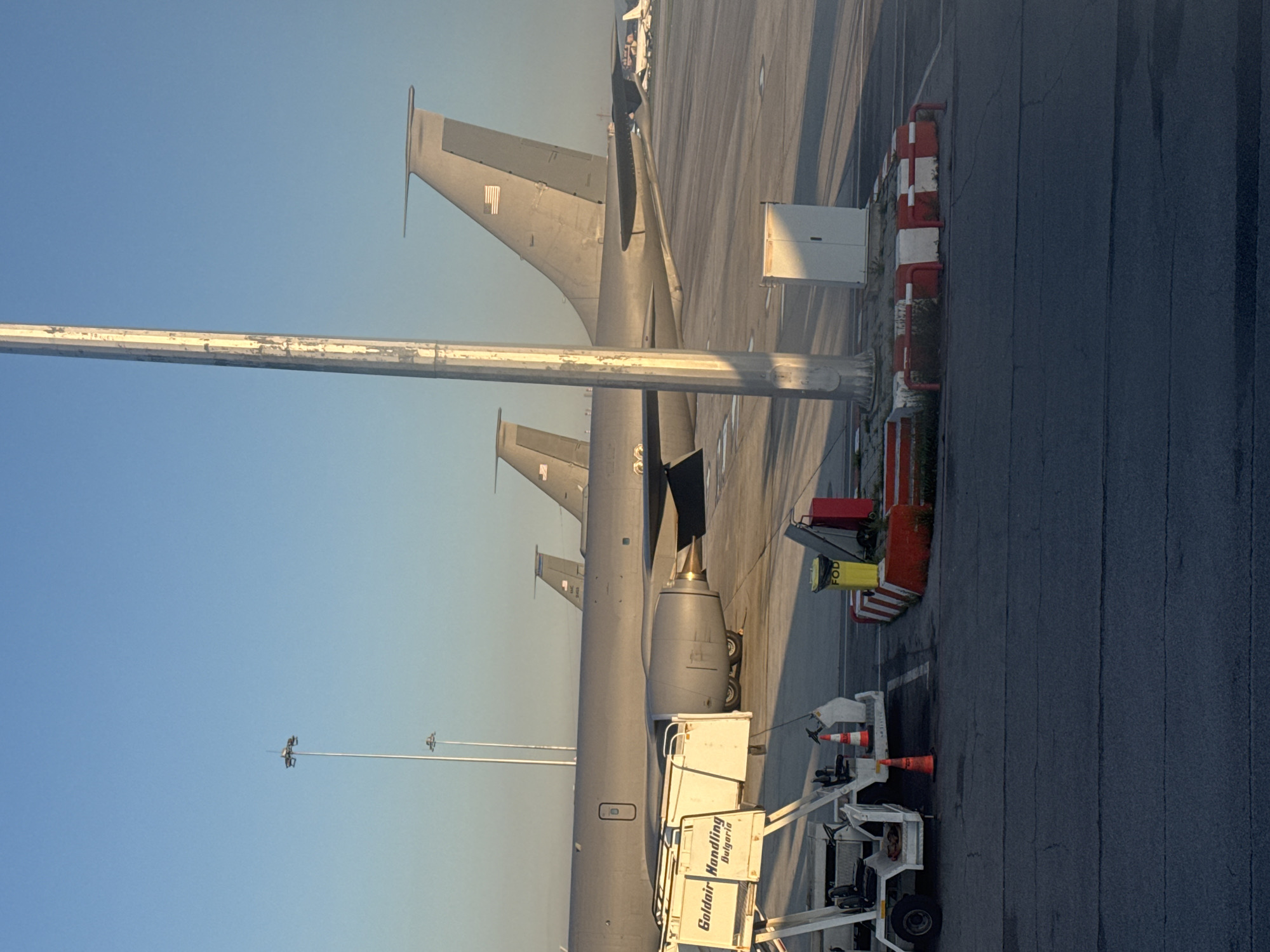The image shows a view of an airport tarmac with several military aircraft lined up, possibly KC-135 Stratotankers, given their tail shape and engine nacelles. A ground service vehicle with "Goldair Handling Bulgaria" is parked in the foreground. Safety cones and barriers are visible alongside the aircraft. The sky is clear and blue. The location is Unknown, Unknown, though the ground handling vehicle implies the scene is in Bulgaria.