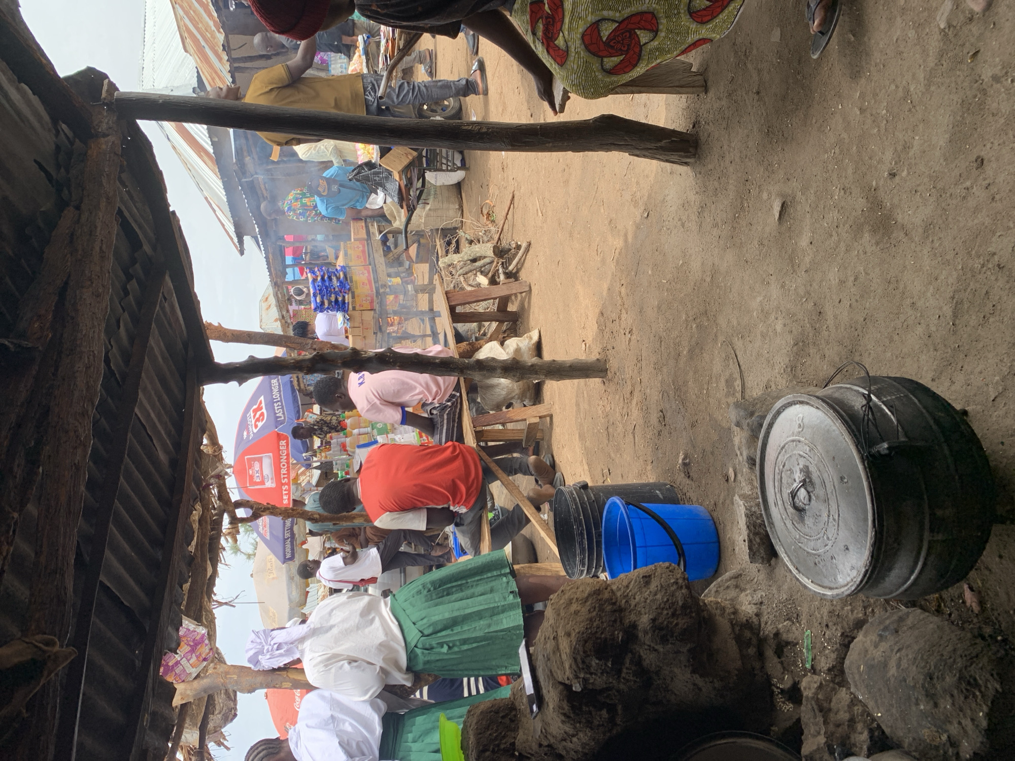 This image depicts a busy market scene in Dull A, Nigeria. Several people, dressed in various casual attire, are visible. Some are seated, others appear to be standing and interacting with each other or the goods on display. The setting is an open-air market with makeshift stalls constructed from corrugated metal and wooden planks. In the foreground, a large, dark pot sits on the ground, suggesting food preparation is underway. Several buckets, one blue and one black, are also present. In the background, more stalls are visible, filled with various items, including what appear to be bottles of liquids and packaged goods.
Smoke is rising from a fire in one of the stalls, indicating cooking is actively happening. The ground is dusty and uneven, typical of an outdoor market. The lighting suggests it is daytime, possibly late morning or afternoon, with no strong shadows indicating harsh sunlight. The overall atmosphere appears active and bustling, with a sense of everyday commerce.
Visible text includes branding on a red and blue umbrella in the background, partially obscured but showing "SETTS STRONGER" and "LASTS LONGER". Another partially visible sign says "Coca-Cola". The scene conveys a sense of community and daily life in a Nigerian market.
