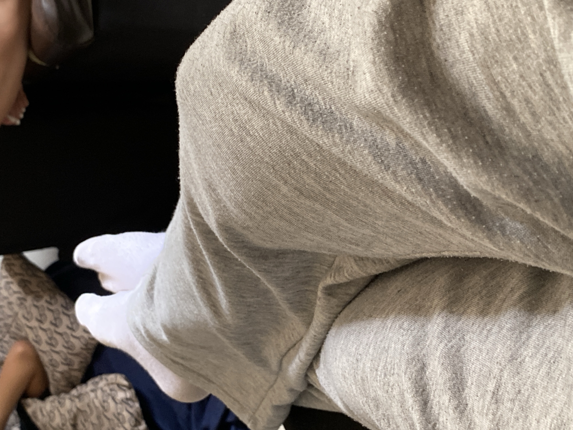 A close-up shot depicts a person's legs, covered in gray sweatpants and white socks. The person is seated on a dark surface, likely a couch or chair. In the lower left corner, a patterned pillow is visible. The location is not discernible from the image.