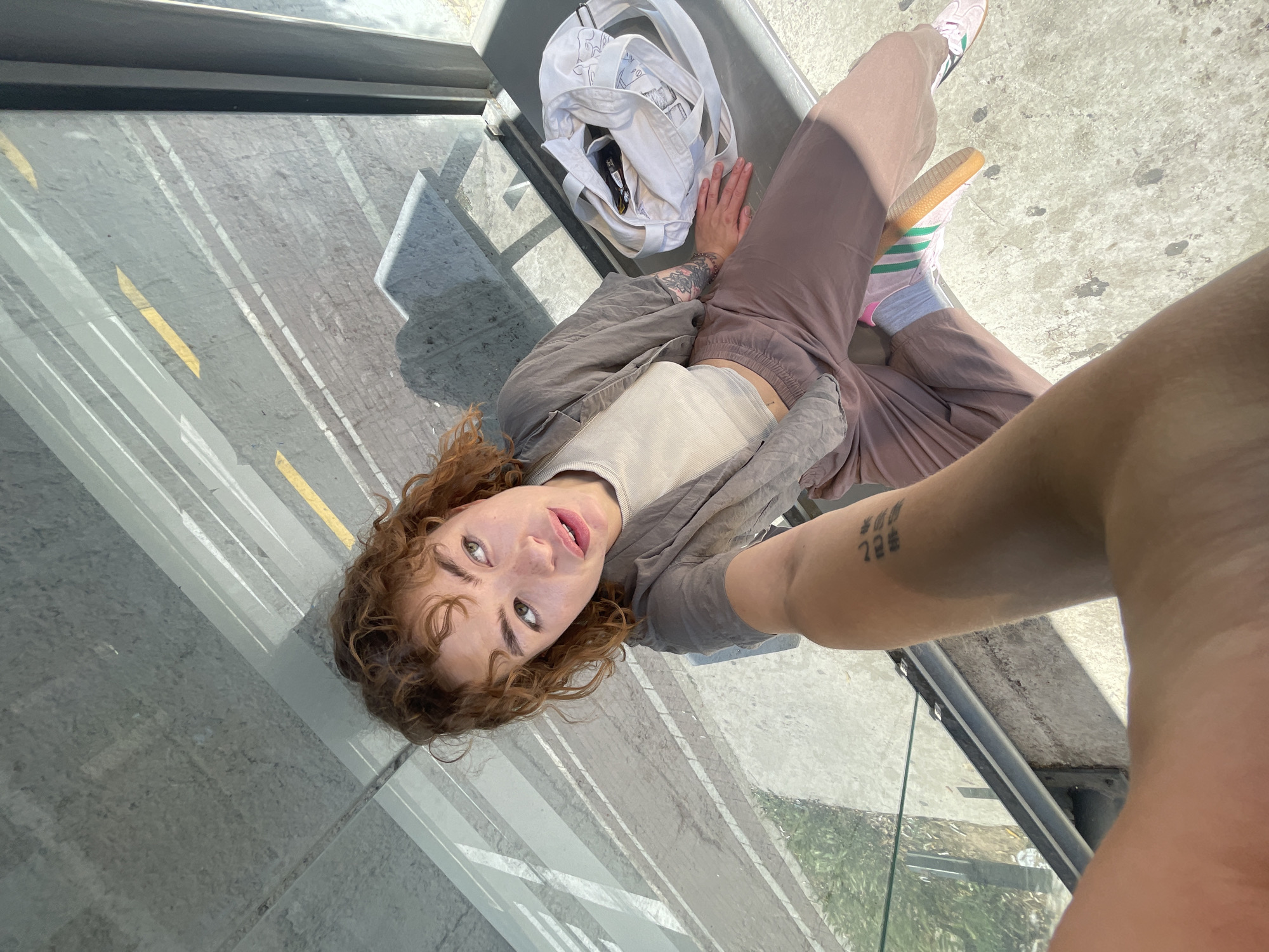 A woman is lying on a public bench in Rafailovići, Montenegro, taking a selfie. She has curly red hair and is wearing brown pants, a light-colored top, and a brown jacket. She has tattoos on both arms. A white tote bag sits on the bench next to her. The background shows a street and building. The image is taken at an extreme angle.