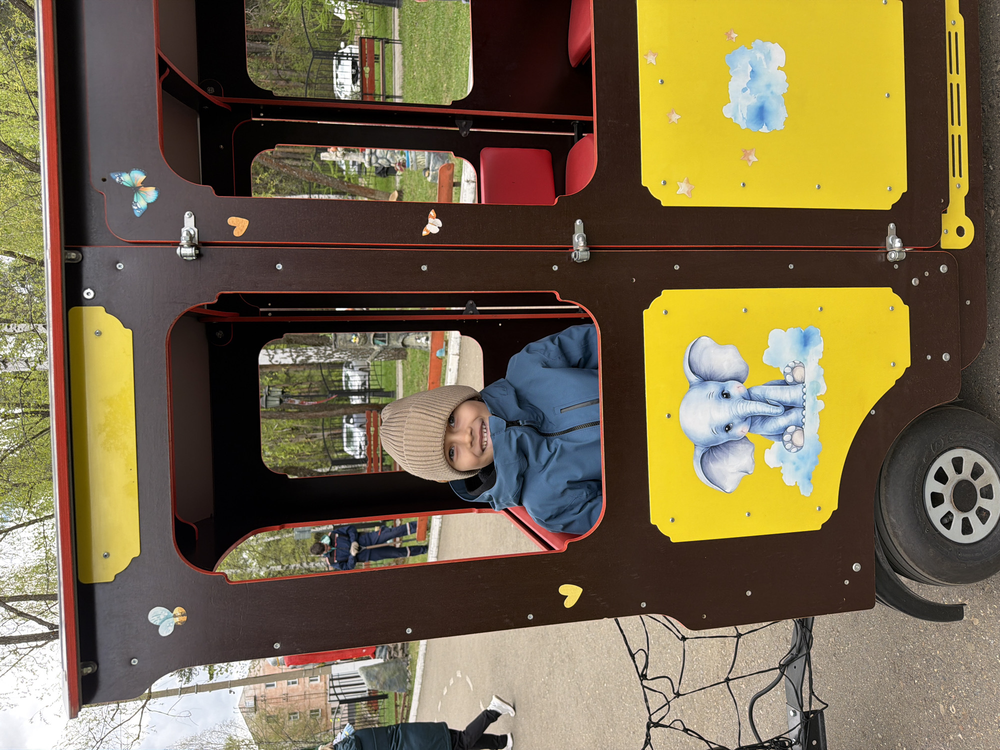 A child in a tan beanie and blue jacket is smiling and looking at the camera from the window of a brown and yellow play structure. The structure has illustrations of elephants and clouds on the yellow panels. It appears to be a stylized train or bus, with a wheel visible on the right side. Behind the structure, the background shows a park-like setting with trees, grass, and building exteriors in the distance. Based on the presence of Russian signs and the user's prompt, this scene is likely located in the city of Chaikovsky, Russia. Two people in uniform can be seen in the middle distance, potentially engaged in yard work.