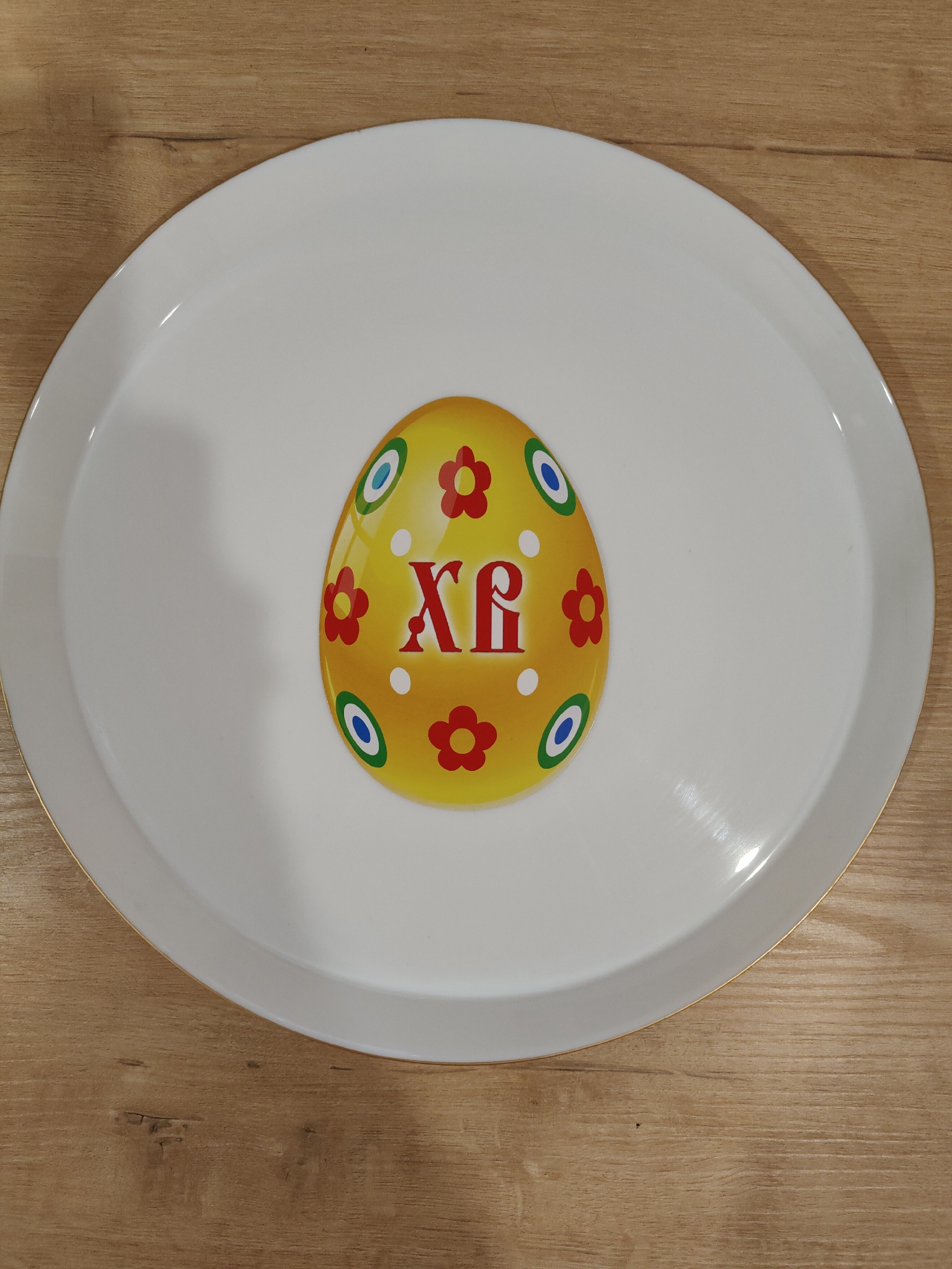 A white oval tray with a gold trim sits on a light brown wooden surface. The tray features a centered, stylized Easter egg design. The egg is yellow with red floral decorations and blue/green circular accents. The letters "XP" are displayed in red in the center of the egg.