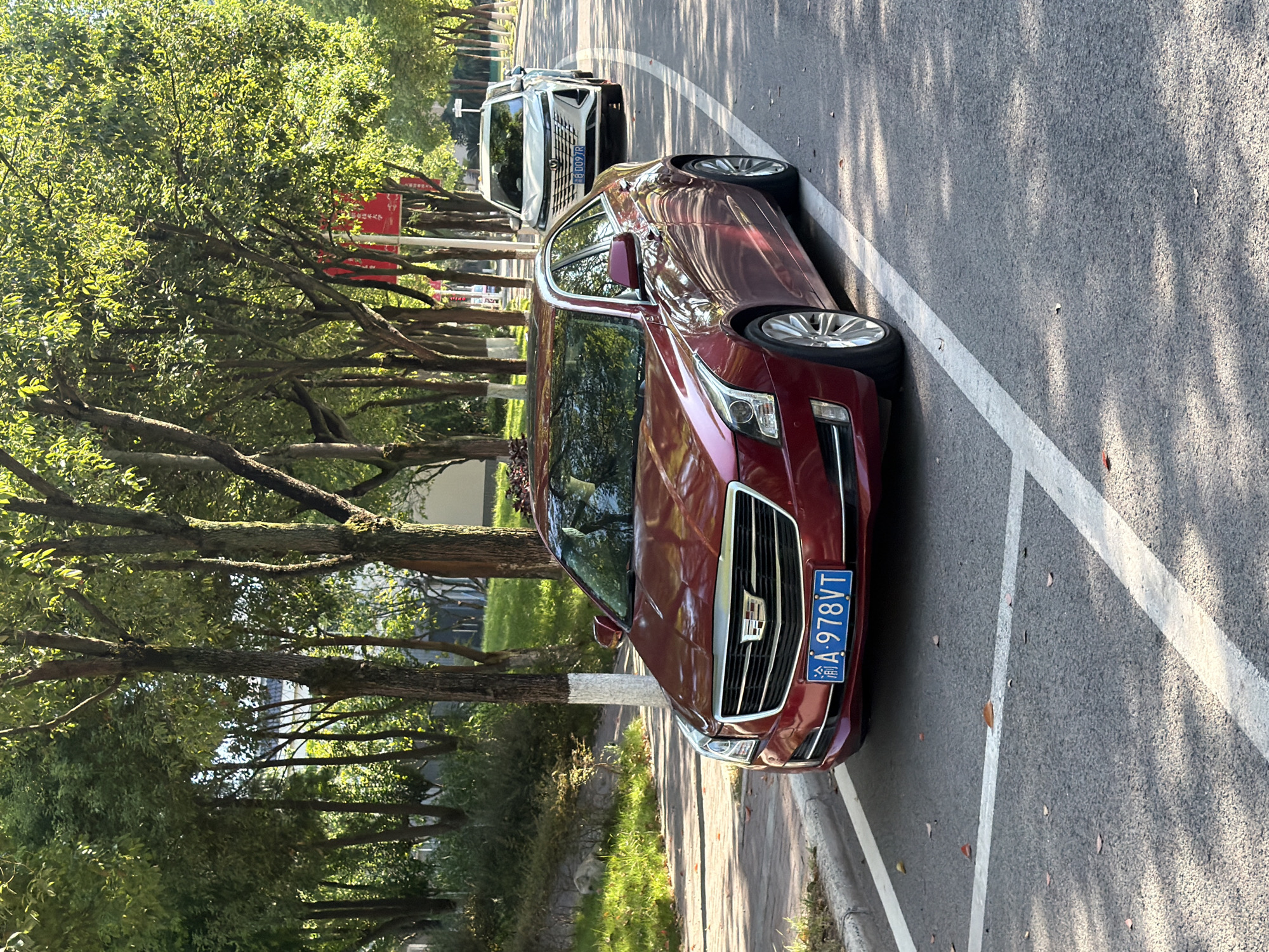 A red Cadillac sedan is parked on the right side of a paved road in Jiangjin, China. The car is facing left, with its front license plate displaying "A-978VT". The road is lined with trees on the left side, casting shadows on the asphalt. A white SUV is parked behind the Cadillac in the same lane. The environment appears to be a suburban or semi-urban street, with a sidewalk and grassy area visible to the left of the Cadillac. The sky is clear and the sunlight suggests it is daytime.