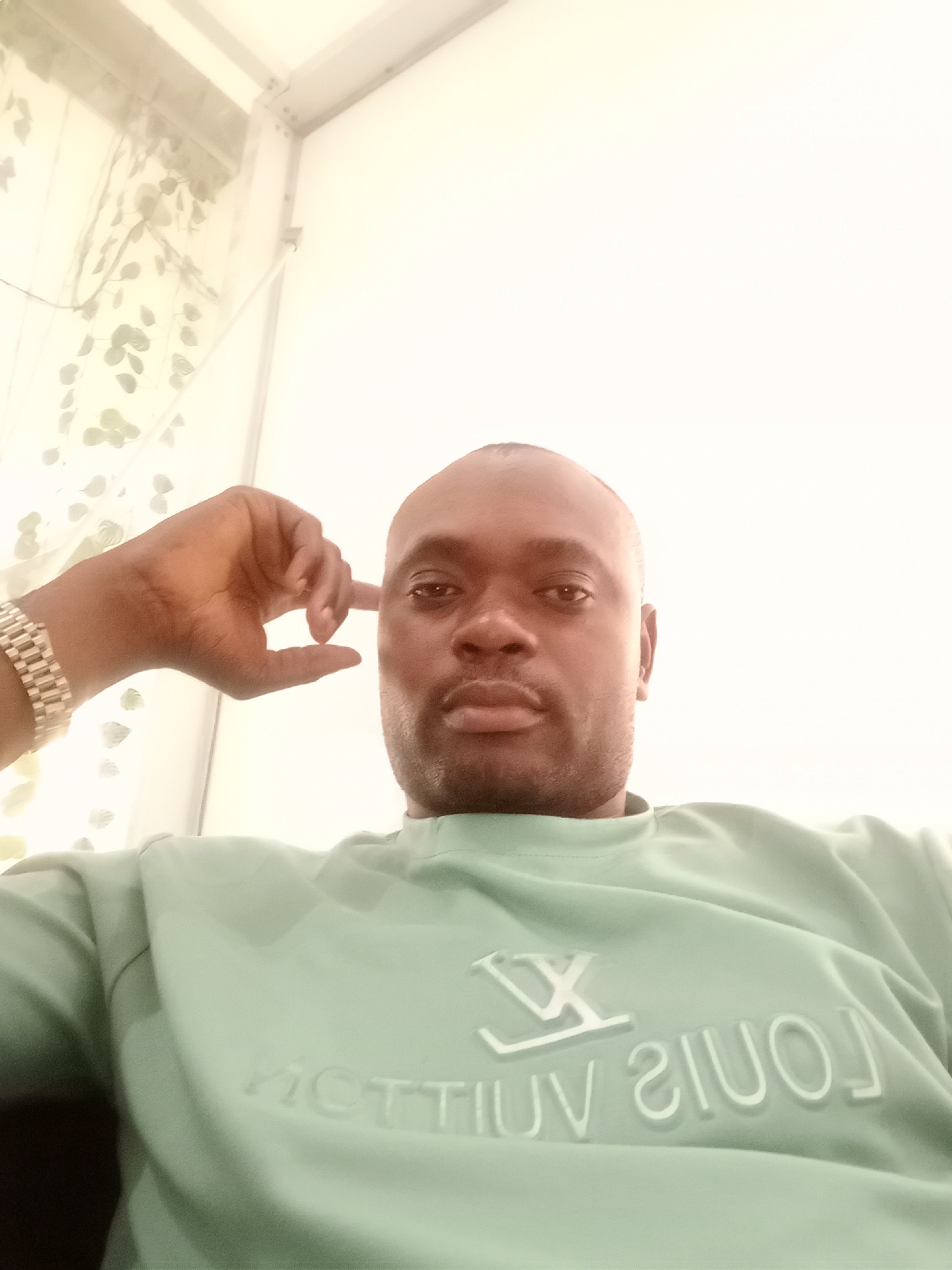 A head-and-shoulders shot from a low angle of a bald man, in Gwarinpa, Nigeria. He is wearing a light green sweater with the letters "LOUIS VUITTON" embossed on the front. The man has dark skin and a neutral expression. His left hand is raised to his head. A silver-colored watch is on his left wrist. The background includes a white wall and a window with sheer curtains featuring a foliage pattern. The image has a slight yellow cast.