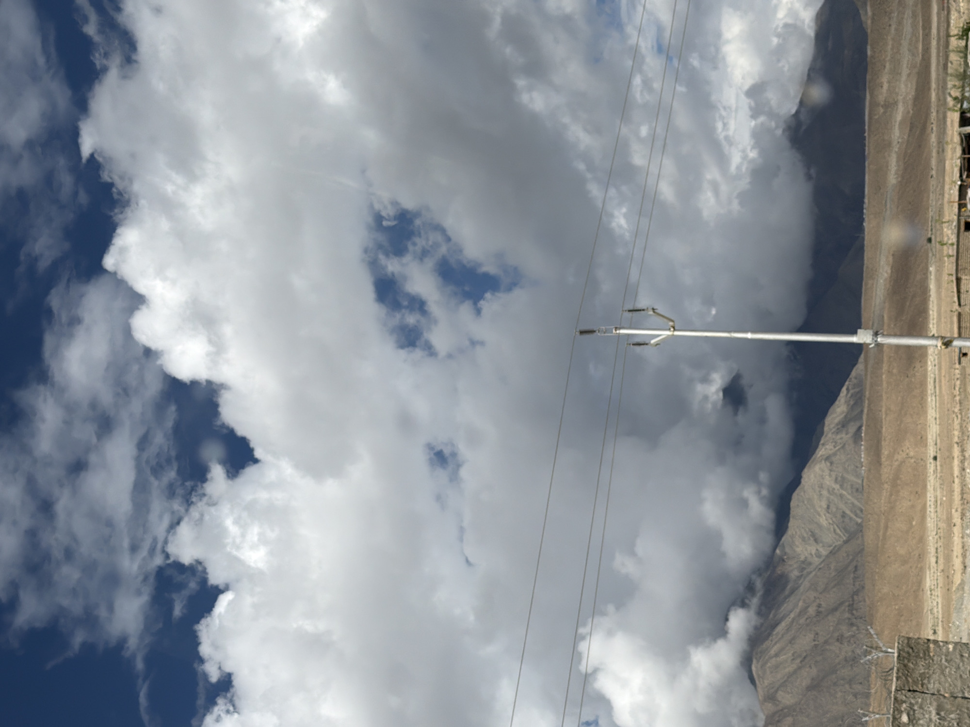 This is a vertical, medium-shot photograph taken from ground level, looking upwards towards a dramatic sky filled with large, fluffy white clouds and patches of deep blue. A single, metallic utility pole with insulators stands prominently in the center of the frame, supporting multiple thin wires that stretch horizontally across the image.
In the background, rugged, barren mountains with discernible slopes and textures are visible. The lower portion of the image shows a dry, desert-like landscape with muted brown and tan tones. A low, rough-textured stone wall or structure is present in the bottom left corner, partially obscuring the ground. In the far distance, there are hints of low-lying buildings or structures. The overall impression is one of a vast, open, and somewhat desolate environment under a dynamic, cloud-filled sky. The location is identified as Chumkar, India.