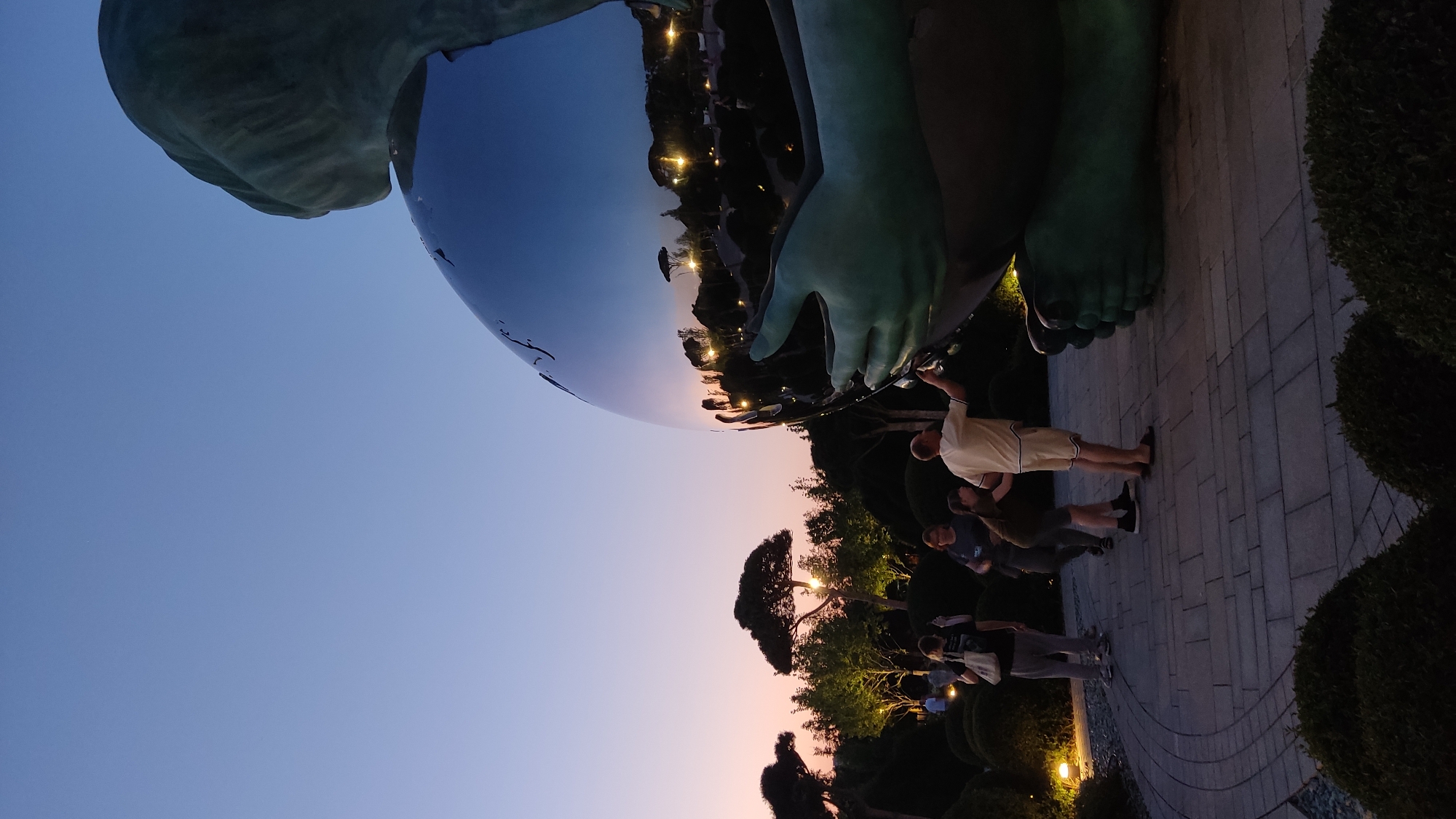 The image depicts a nighttime scene at an outdoor location, likely a park or garden, in Krasnodar, Russia. The focal point is a large, highly reflective metallic sculpture, possibly depicting a figure or a globe, which mirrors the surrounding environment.
Several people are visible in the foreground and midground, interacting with the sculpture and the space. One man in a light-colored t-shirt and shorts stands near the base of the sculpture, appearing to pose for a photo. Another person is partially visible behind him. Further in the midground, a group of individuals are gathered, some looking at the sculpture, others conversing. The figures are silhouetted against the dim lighting and the twilight sky.
The setting includes manicured bushes and trees, a paved walkway with a curved design, and what appear to be accent lights illuminating the foliage and pathway. The sky exhibits the soft hues of twilight, transitioning from a pale blue at the top to a gradient of pink and orange near the horizon, indicating the time of day is dusk or early evening. The reflections in the sculpture show lights from the park, trees, and the sky.
The overall mood is relaxed and social, suggesting people are enjoying an evening out. No visible text is present in the image. The metallic sculpture dominates a significant portion of the frame, its polished surface capturing a distorted yet recognizable reflection of the scene.
