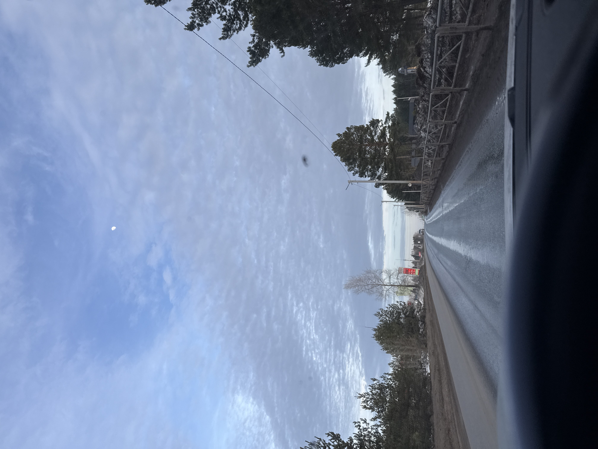 The image depicts a forward view from inside a vehicle traveling on a two-lane asphalt road in Chaikovsky, Russia. The sky is largely covered by scattered altocumulus clouds with patches of pale blue, and a visible crescent moon is positioned in the upper center-right. A dark, circular smudge is present on the upper right portion of the visible sky, likely on the vehicle's windshield.
The road surface appears wet or damp, reflecting the sky in places, and has a visible, faded double white line marking the center, extending into the distant horizon. The shoulders of the road are unpaved, consisting of dark earth and patches of accumulated snow or slush.
On the left side of the road, a dense line of coniferous trees forms a forest edge. Further along, a bare deciduous tree with light bark is visible, alongside a red vertical sign displaying text including "AHTE" and "P-172". Utility poles with power lines run parallel to the road on this side.
On the right side of the road, a weathered wooden fence with a diagonal cross-hatch design runs adjacent to the road. Behind the fence are accumulations of dirt and snow. Various coniferous trees, some quite large, are present, partially obscuring distant structures, including a dark-roofed building and a light-colored tower-like object. Additional utility poles and overhead wires are visible on this side, with one prominent wire stretching diagonally across the sky from a pole on the right.
The bottom approximately 15% of the image shows the dark, unidentifiable dashboard or interior surface of the vehicle. No individuals or specific actions are discernible within the frame.