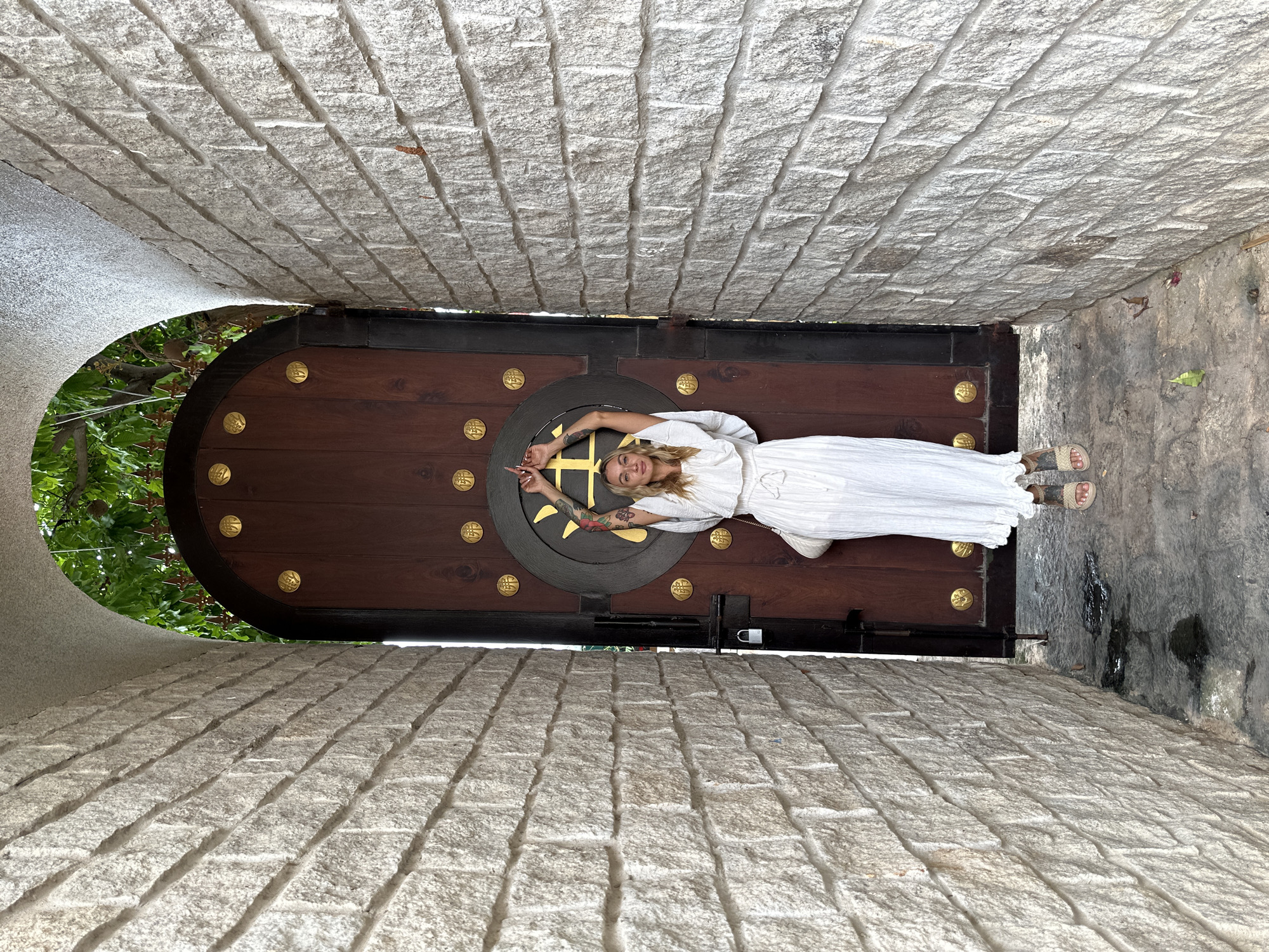 The image shows a woman lying on a dark wooden door with ornate gold embellishments, set within a narrow stone passageway in Mũi Né, Vietnam. The woman is wearing a white dress and sandals, and has visible tattoos on her arm. The door is large and imposing, with a circular central design. The passageway walls are constructed of light-colored, rough-hewn stone blocks. A glimpse of green foliage is visible through an archway to the left. The scene is taken from a low angle, making the passageway seem elongated.